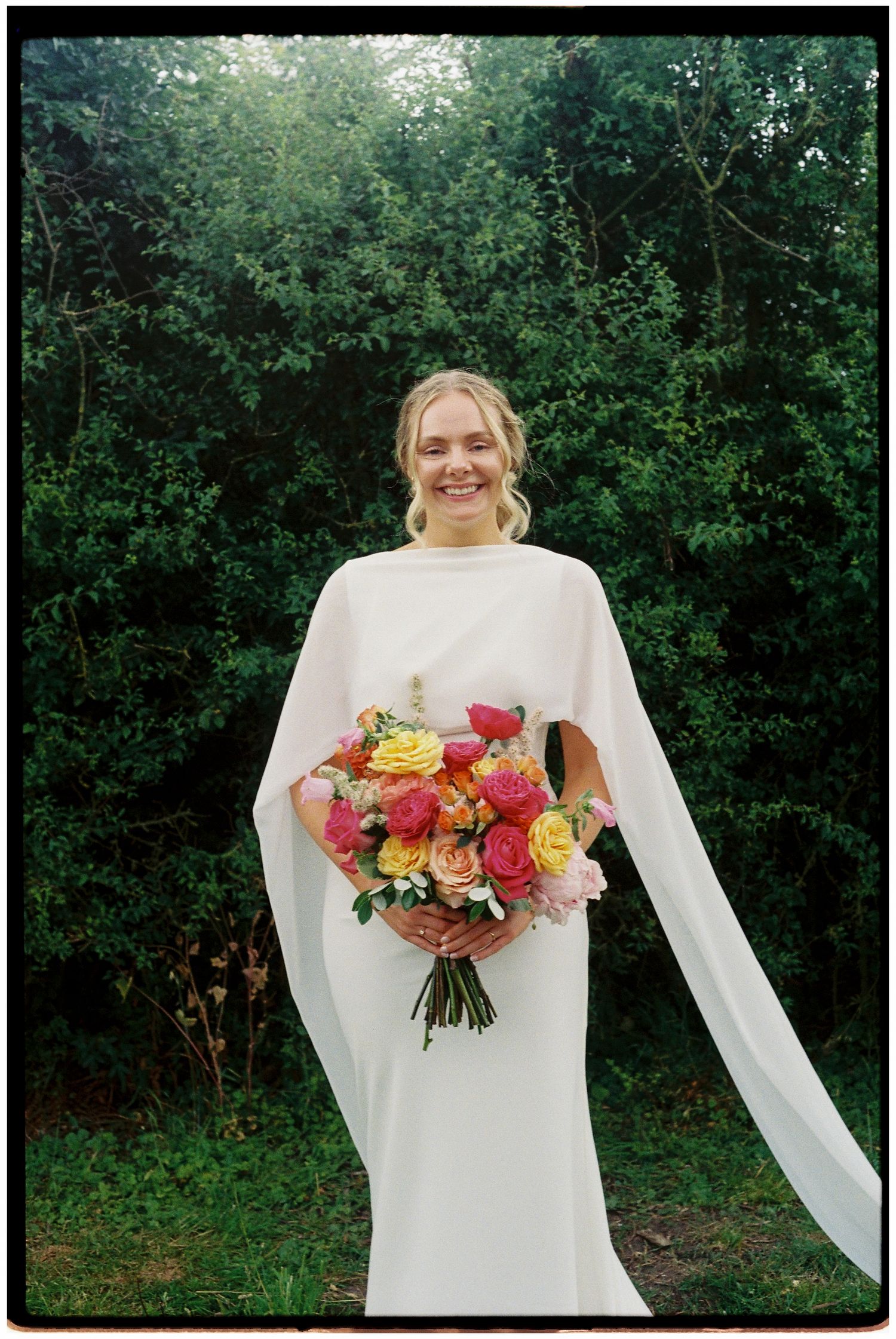 Smiling bride in elegant white cape gown holds a colorful bouquet of roses outdoors.