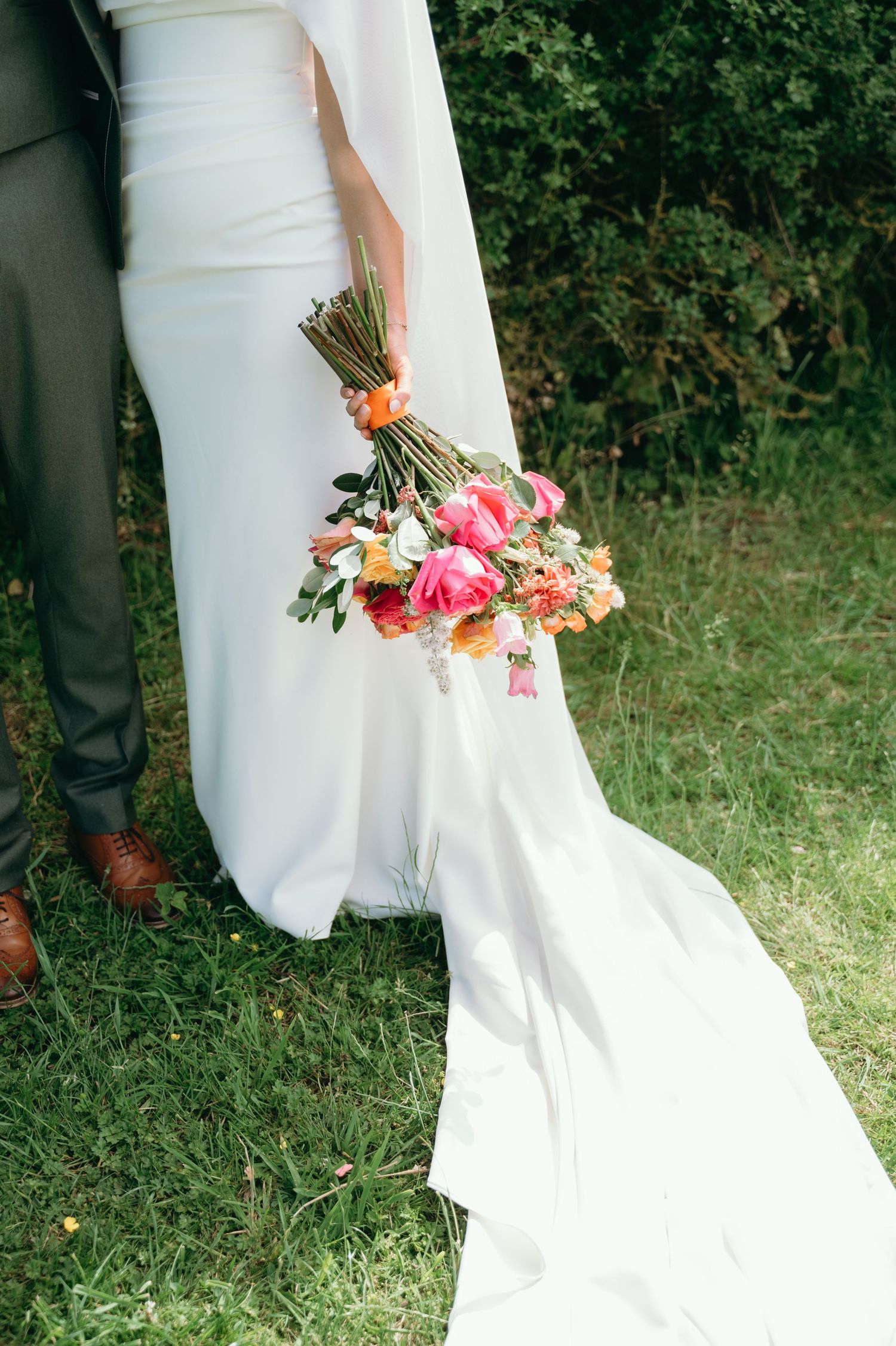 Bride in white gown with train holds a vibrant pink and orange bouquet beside her groom.