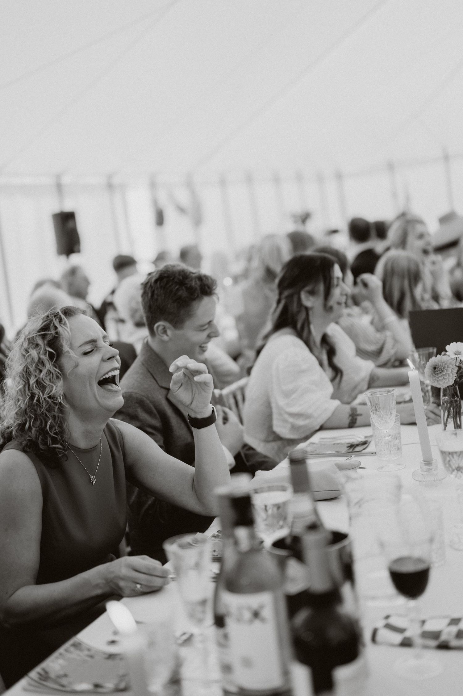 Black and white photo of wedding guests laughing joyfully at long reception tables inside a marquee tent.