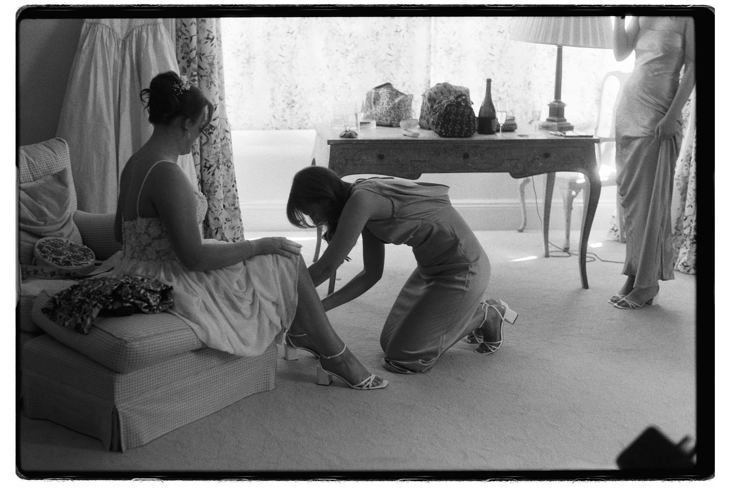 Black and white film photo of a bridesmaid kneeling to fasten another womans sandal in an elegant room.