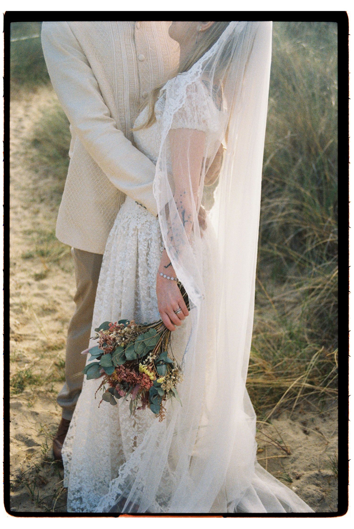 Bride in lace gown and veil holds bouquet while embracing groom on sandy dunes.