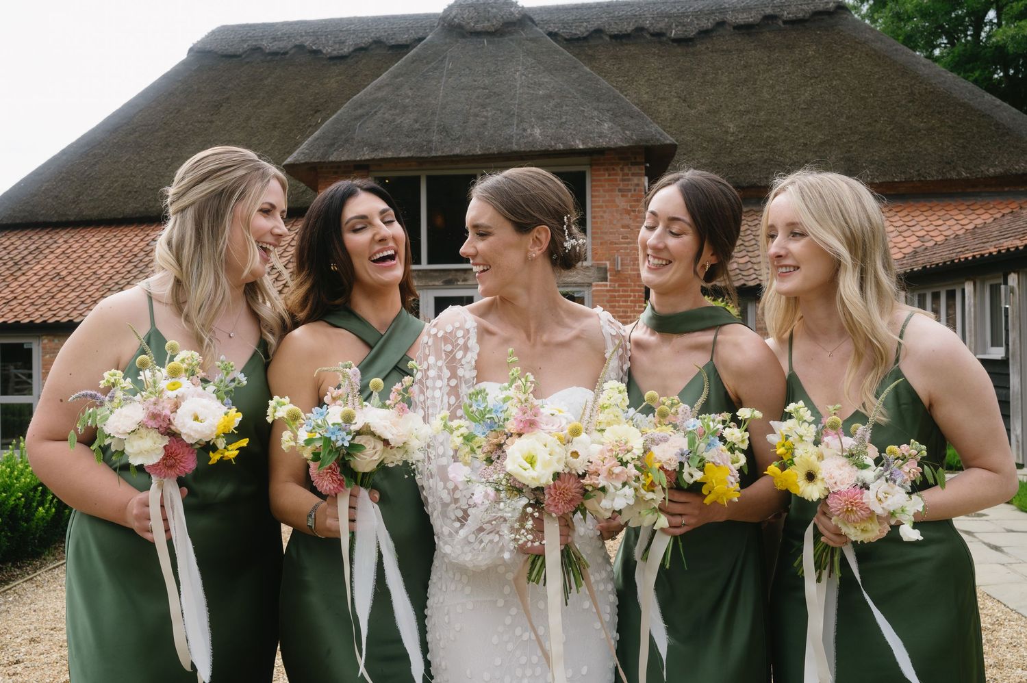 Bride with four bridesmaids in sage green dresses holding floral bouquets outside barn venue.