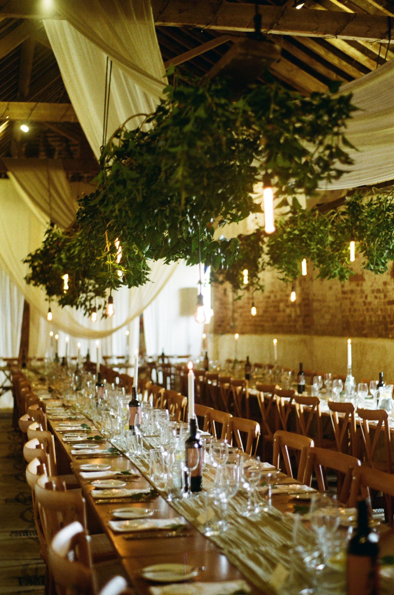 Rustic barn wedding reception with long tables, candles, wine bottles, and lush hanging greenery ceiling.