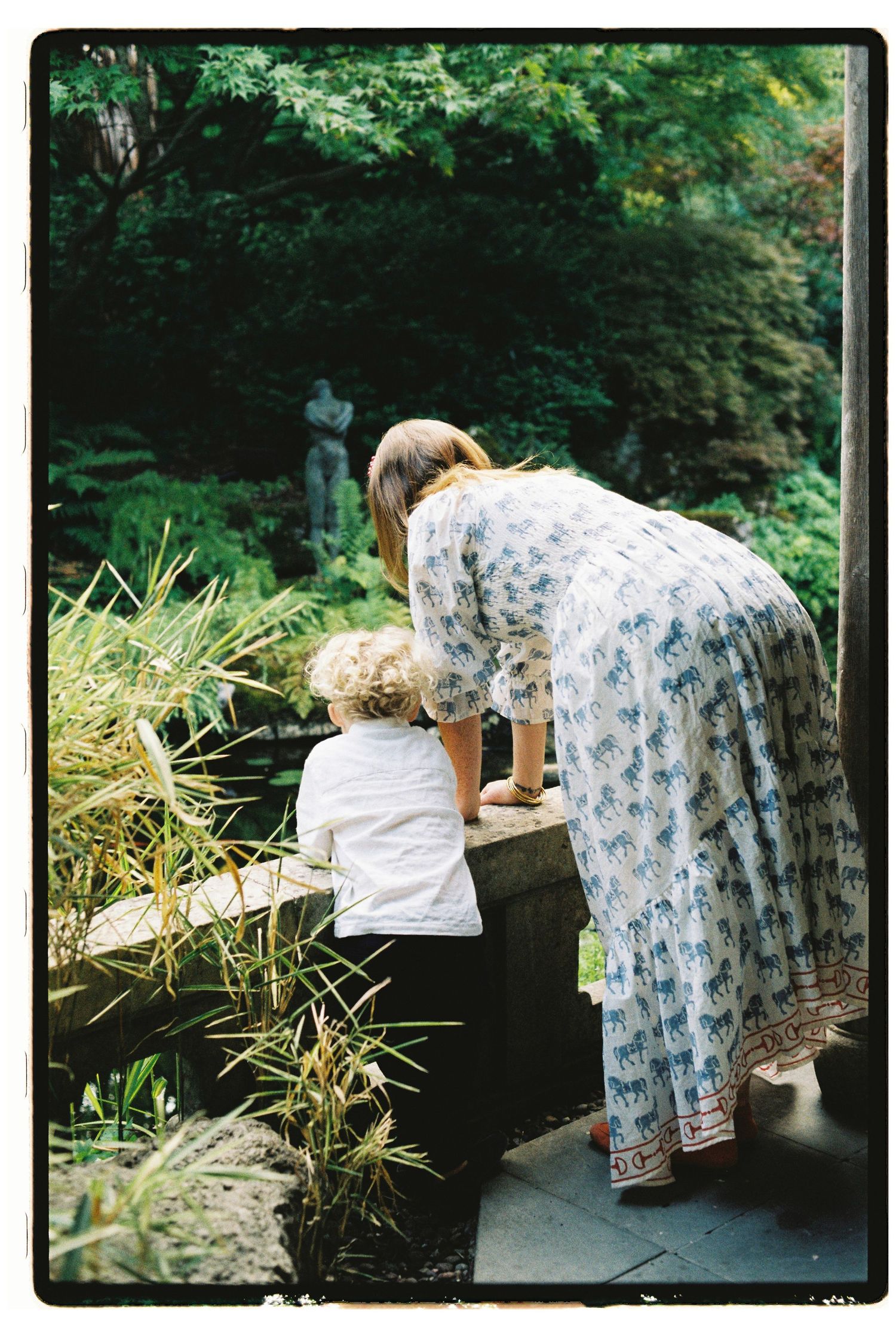 Color film photo of a woman in a floral dress bending over a stone wall with a young child in a garden.