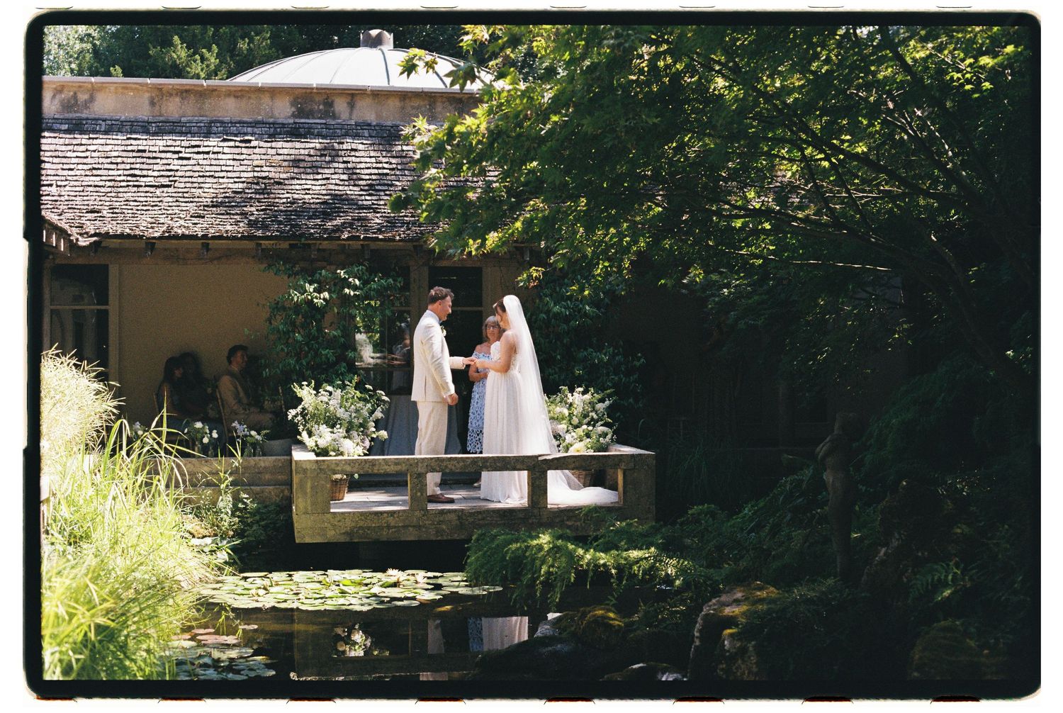 Bride and groom exchanging vows on a wooden bridge surrounded by lush garden and lily pond.