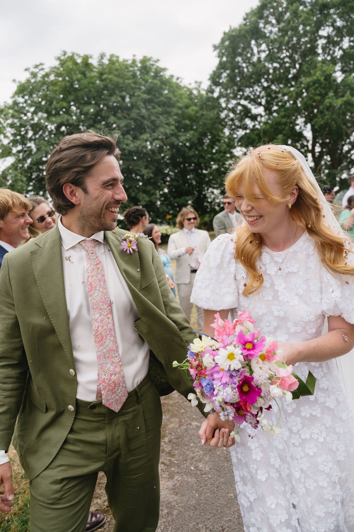 Laughing bride in white floral dress and groom in green suit walk together after ceremony.