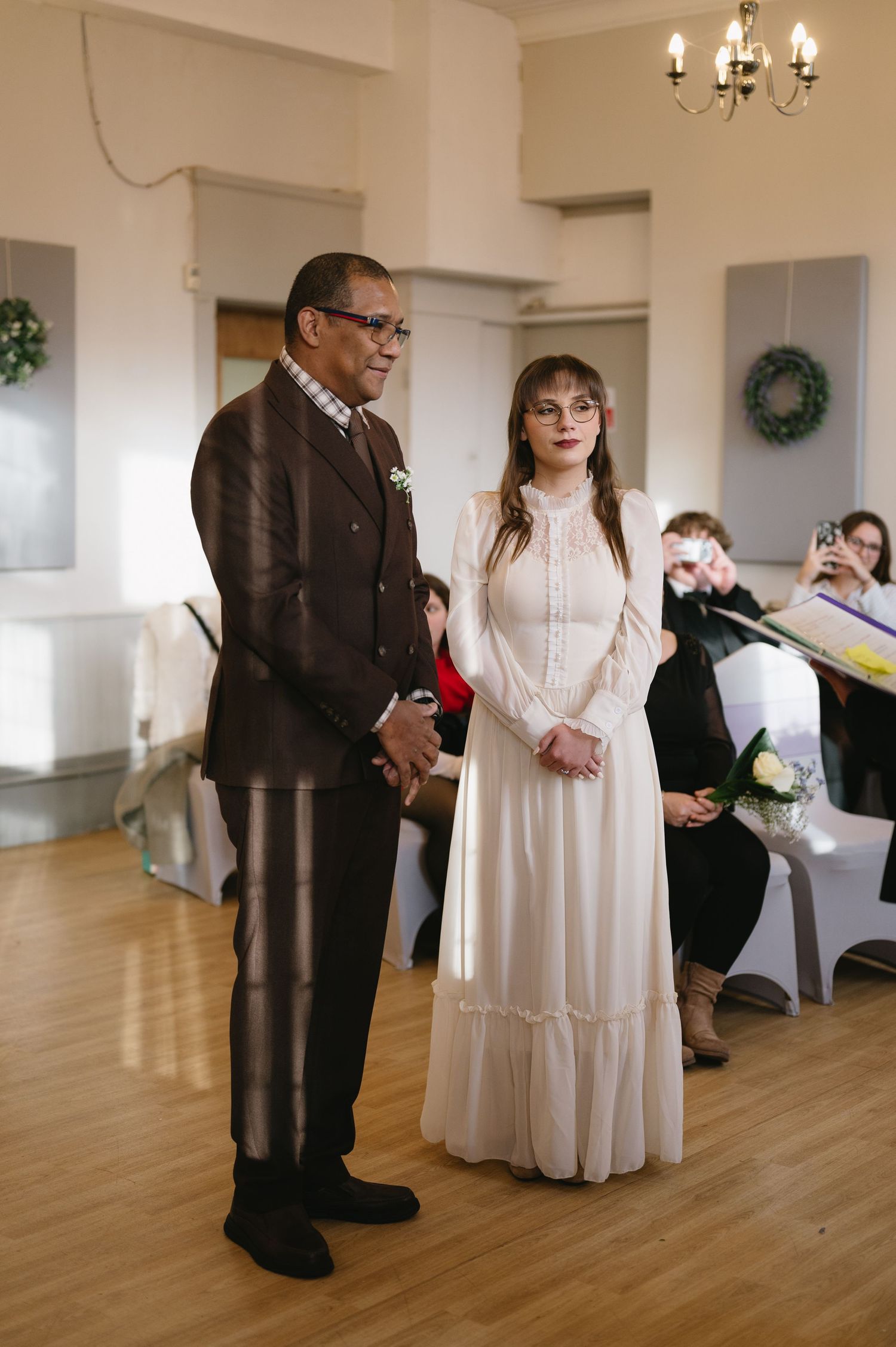 couple stand together during indoor wedding ceremony in vintage-style white gown and suit.