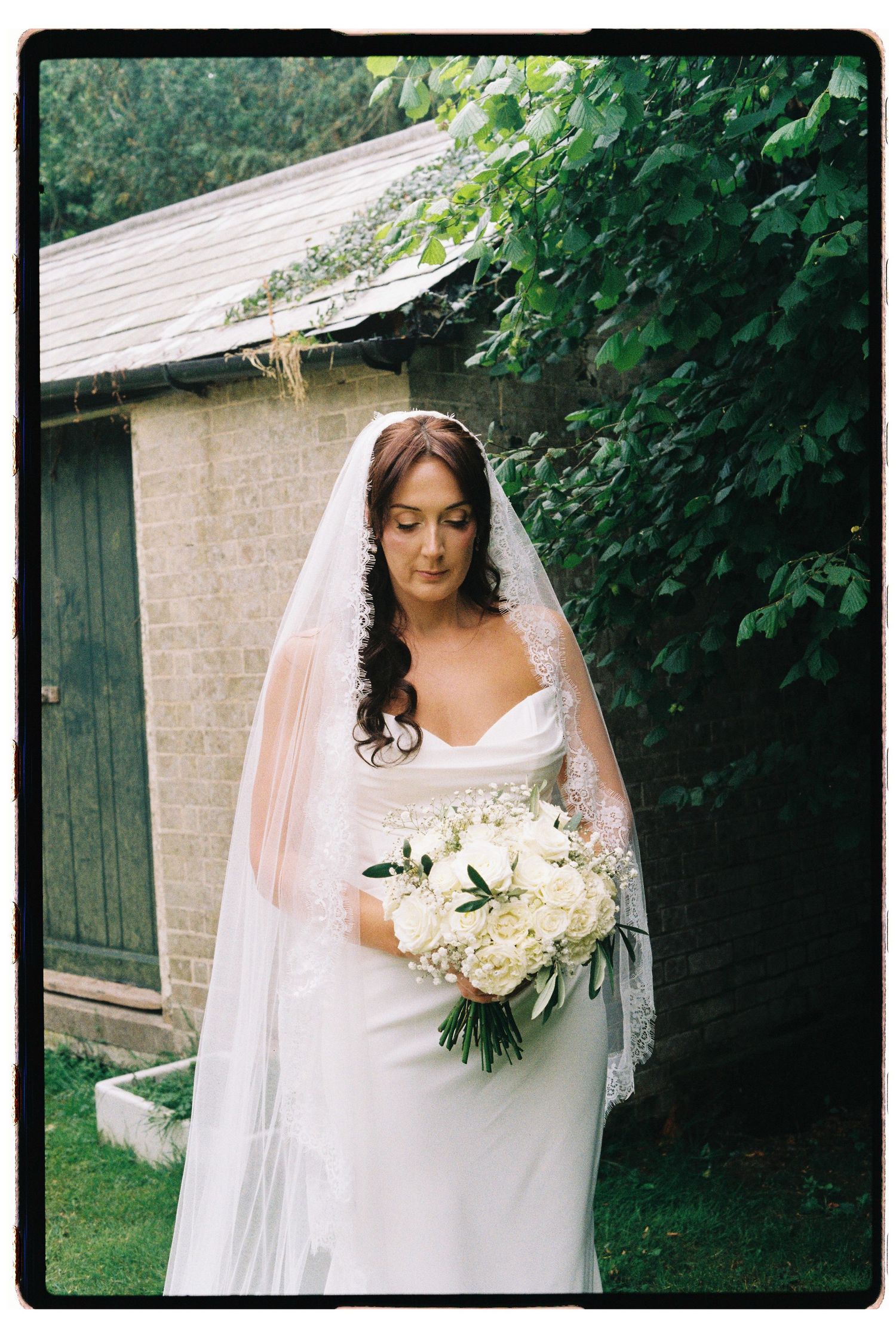 Elegant bride in fitted white gown and lace veil holds white rose bouquet outdoors.