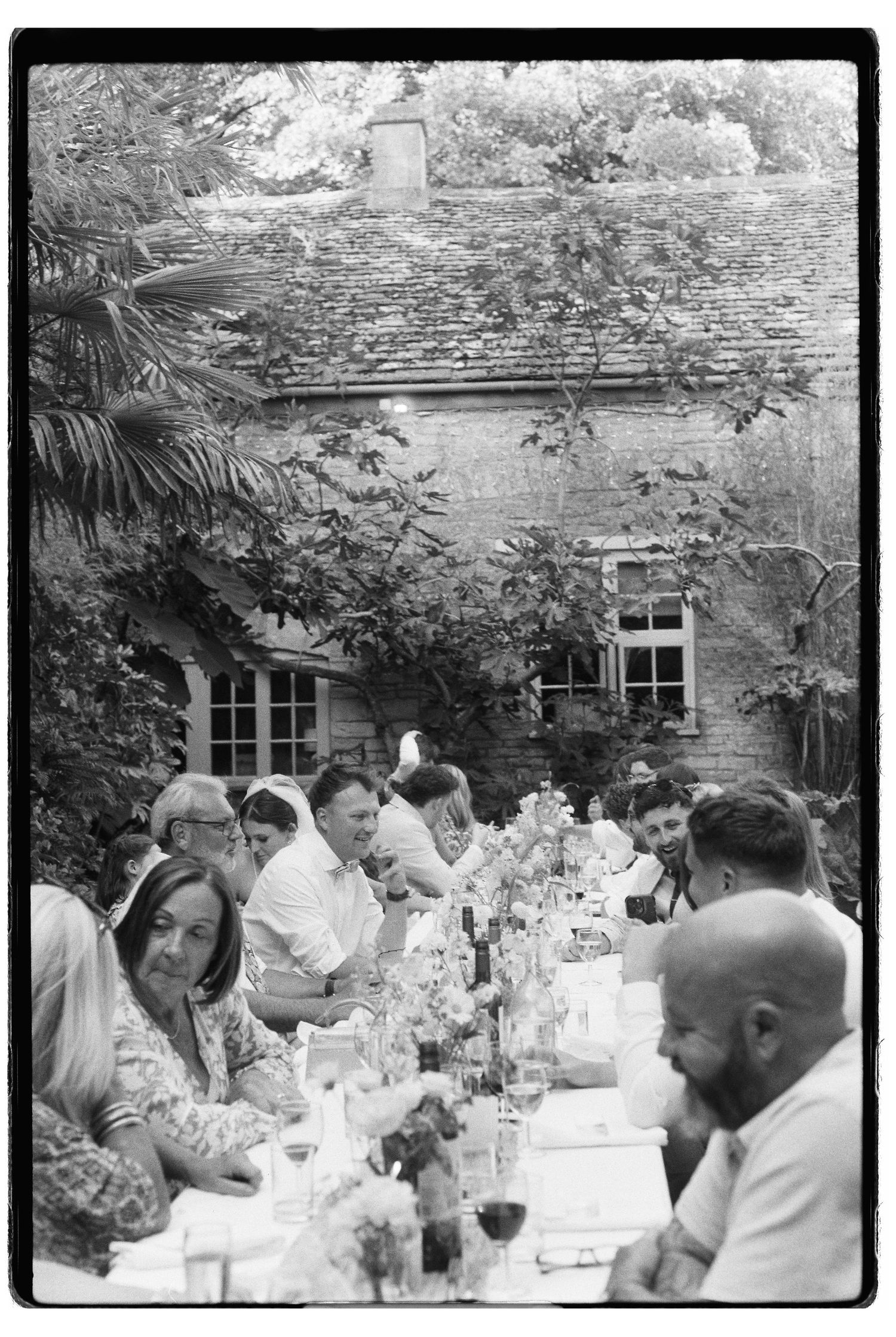 Black and white film photo of wedding guests dining outdoors at long tables beside a rustic stone building.