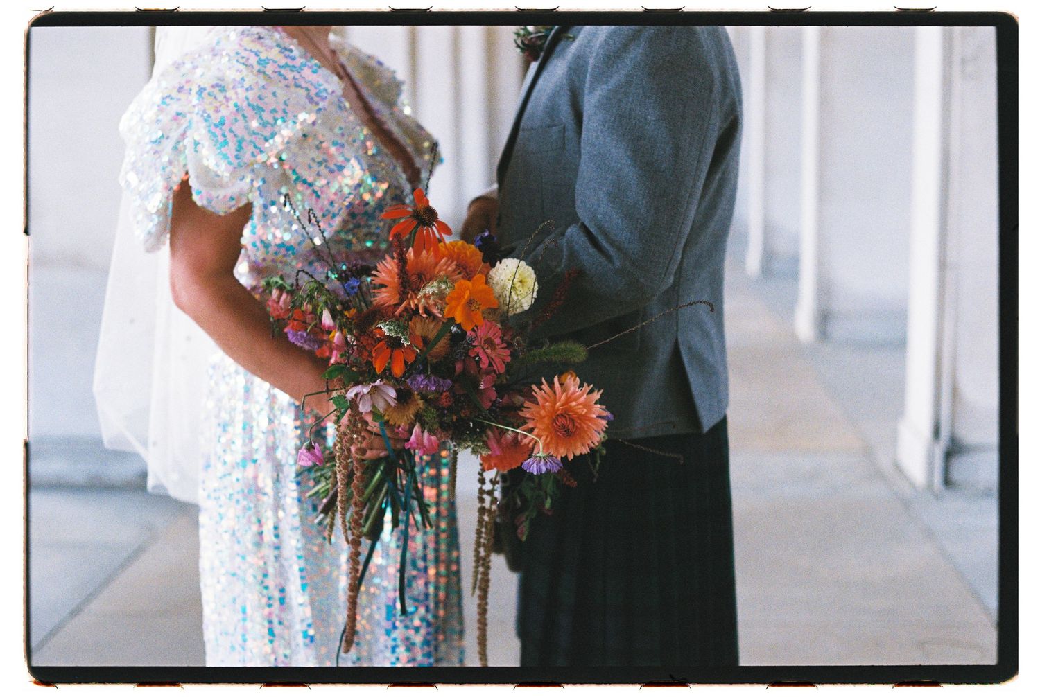 Color film photo of a couple holding a vibrant bouquet of orange and pink flowers at their wedding.