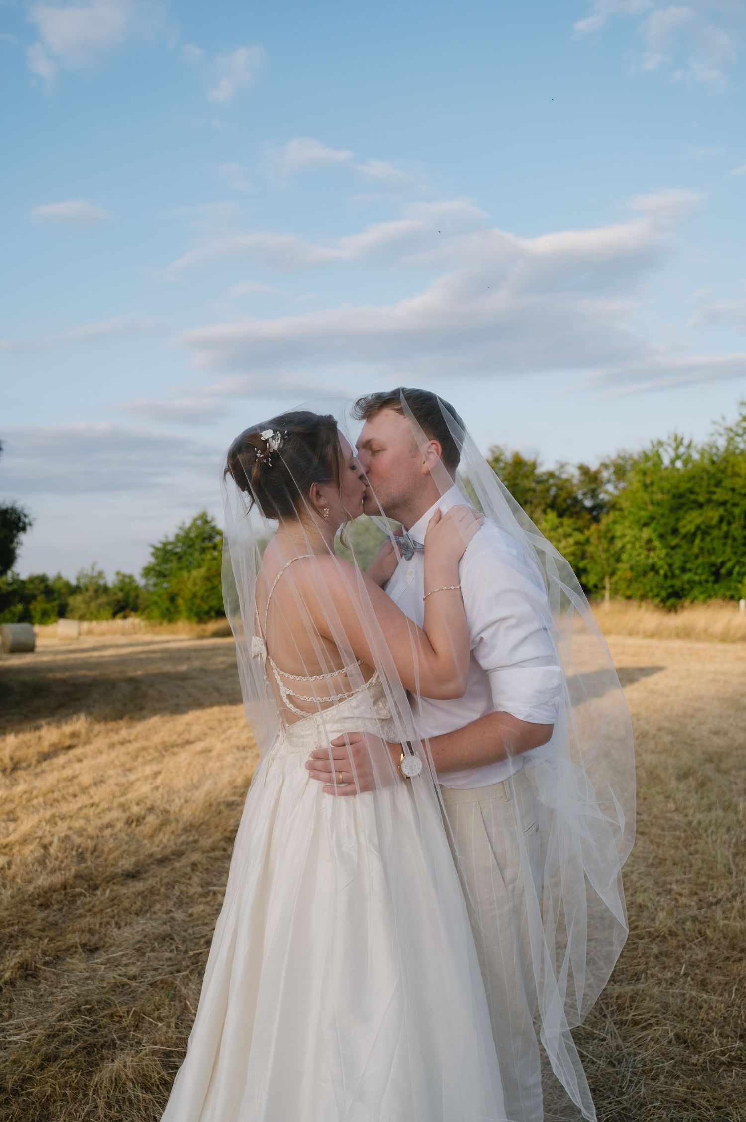 Bride and groom kissing under a veil in a golden field at sunset on their wedding day.
