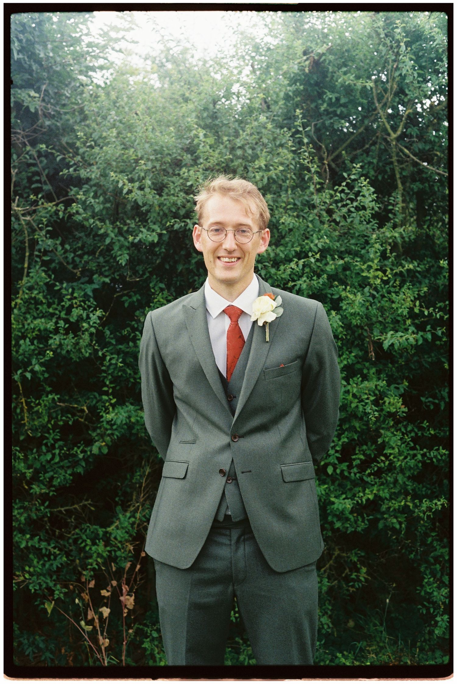 Smiling groom in grey suit with red tie and boutonniere stands before lush green hedges.