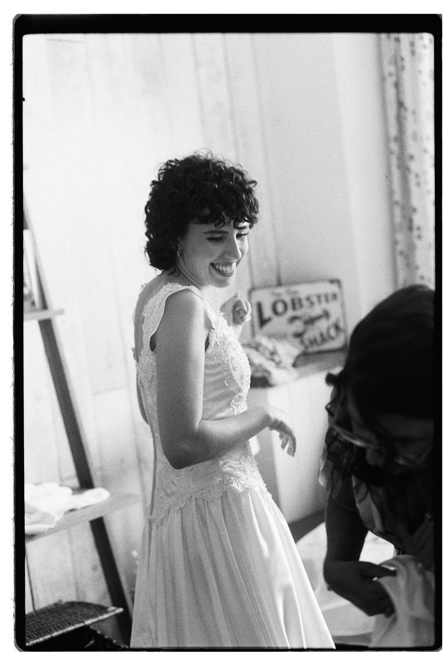 Bride in white lace wedding dress laughing joyfully while being helped by bridesmaid in black and white photo.