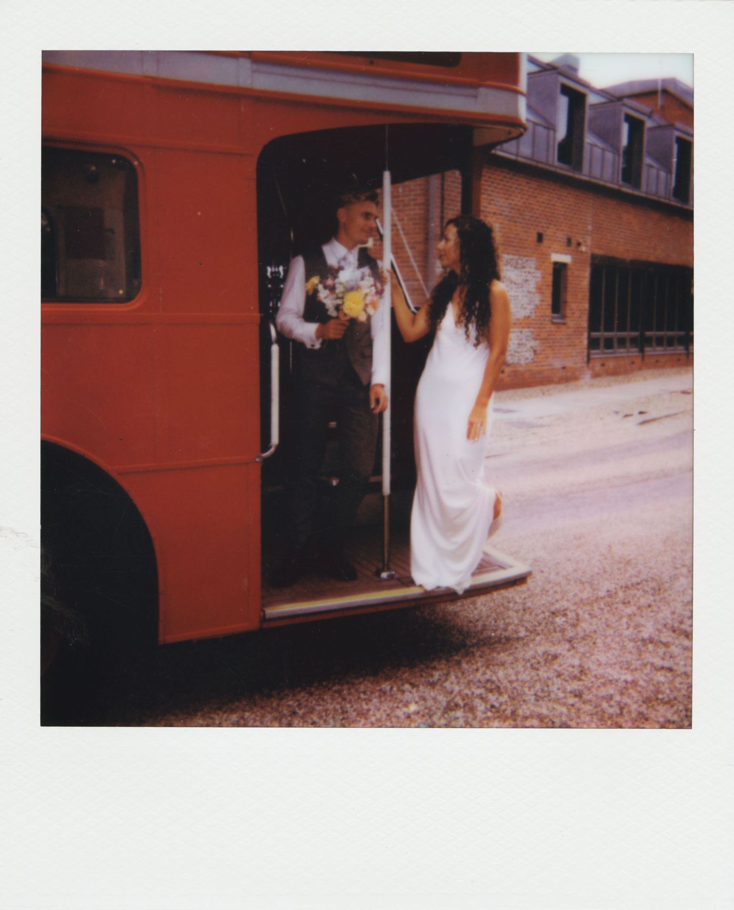 Bride in white dress and groom with bouquet stepping off a vintage red bus on their wedding day.