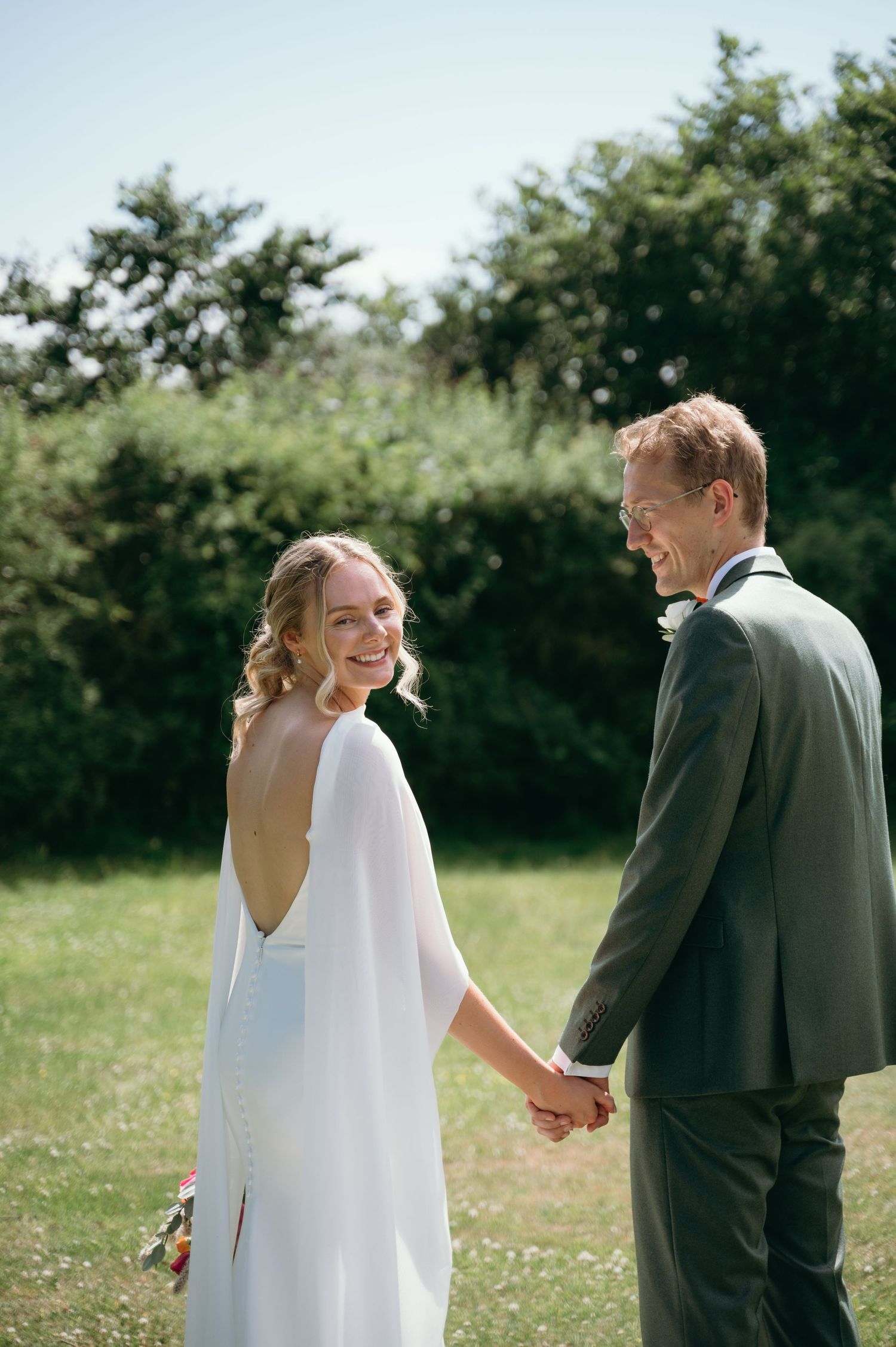 Smiling bride in backless white cape dress holds hands with groom in grey suit outdoors.