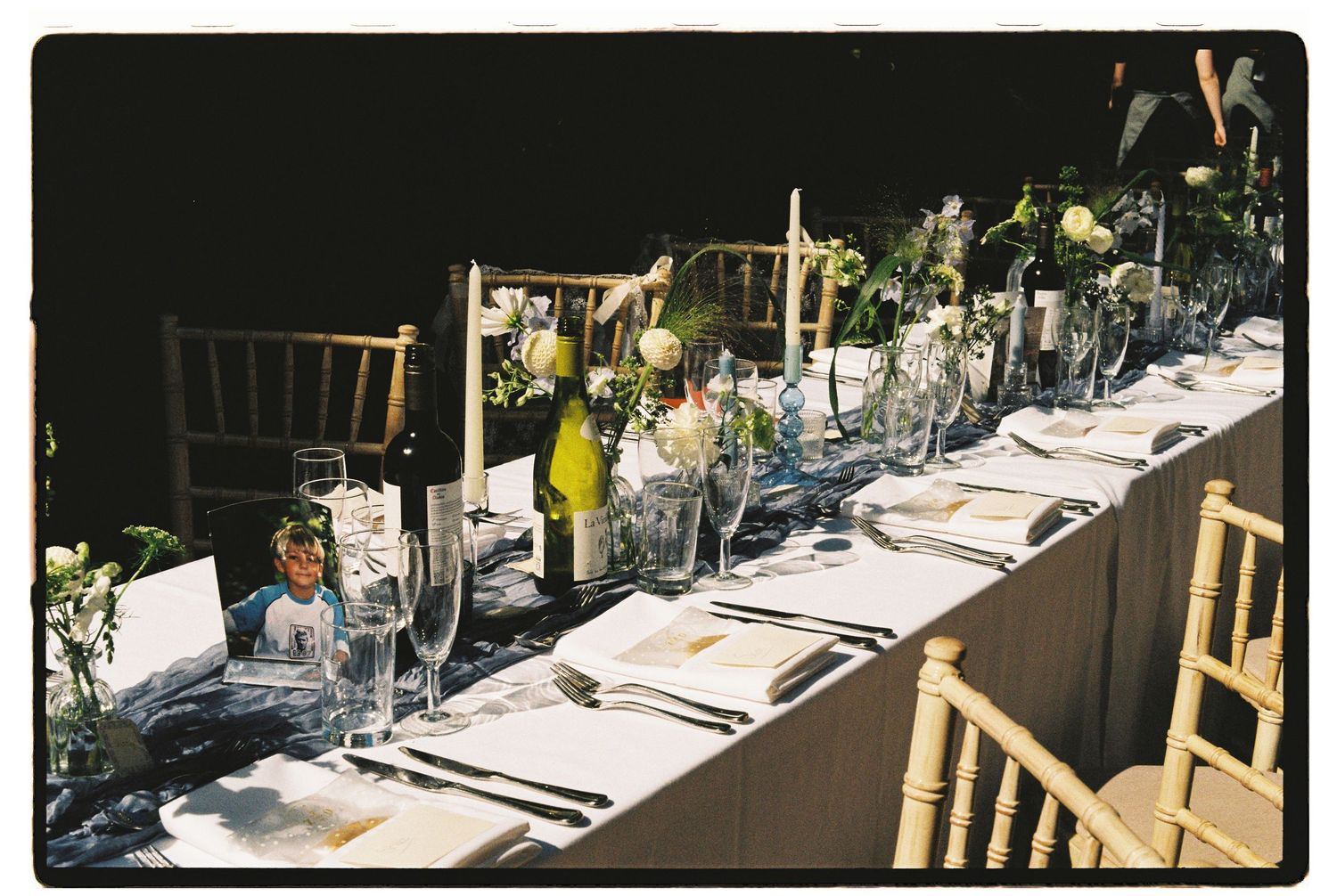 Elegant long wedding reception table set with glassware, flowers, and candles in dim lighting.