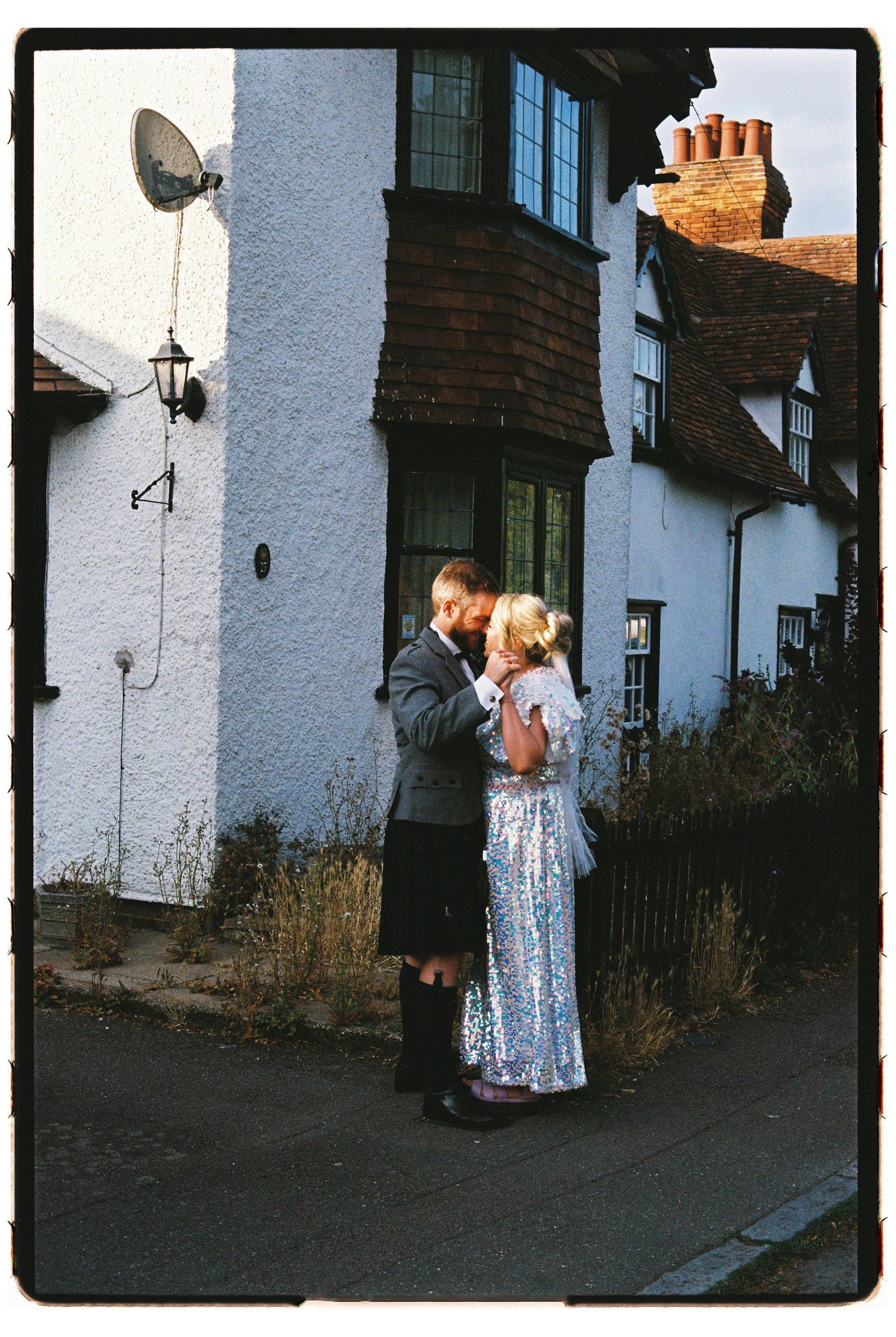 Bride in sequin dress and groom in kilt share a tender moment outside a white cottage.