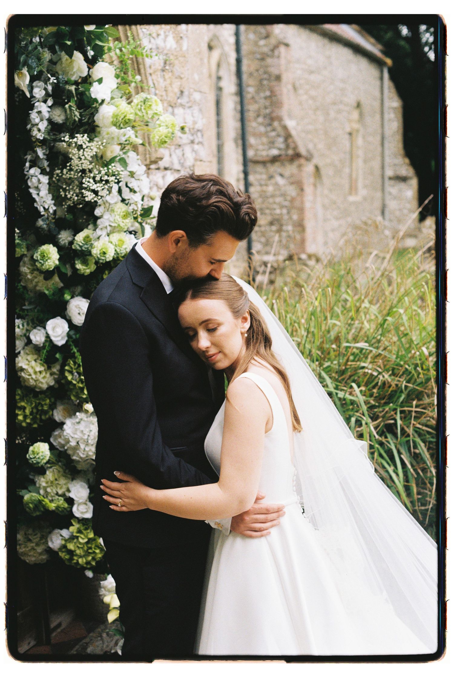 Bride in white ballgown and groom in black suit embrace tenderly beside a floral arch at a stone church.
