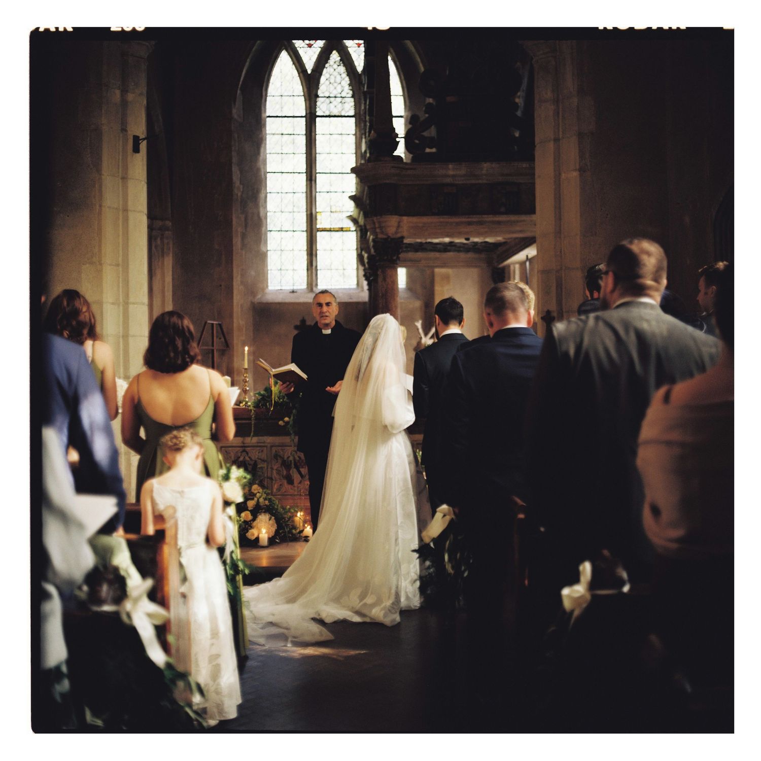 Bride in a long white gown and veil stands before an officiant during a candlelit wedding ceremony in a historic stone church.