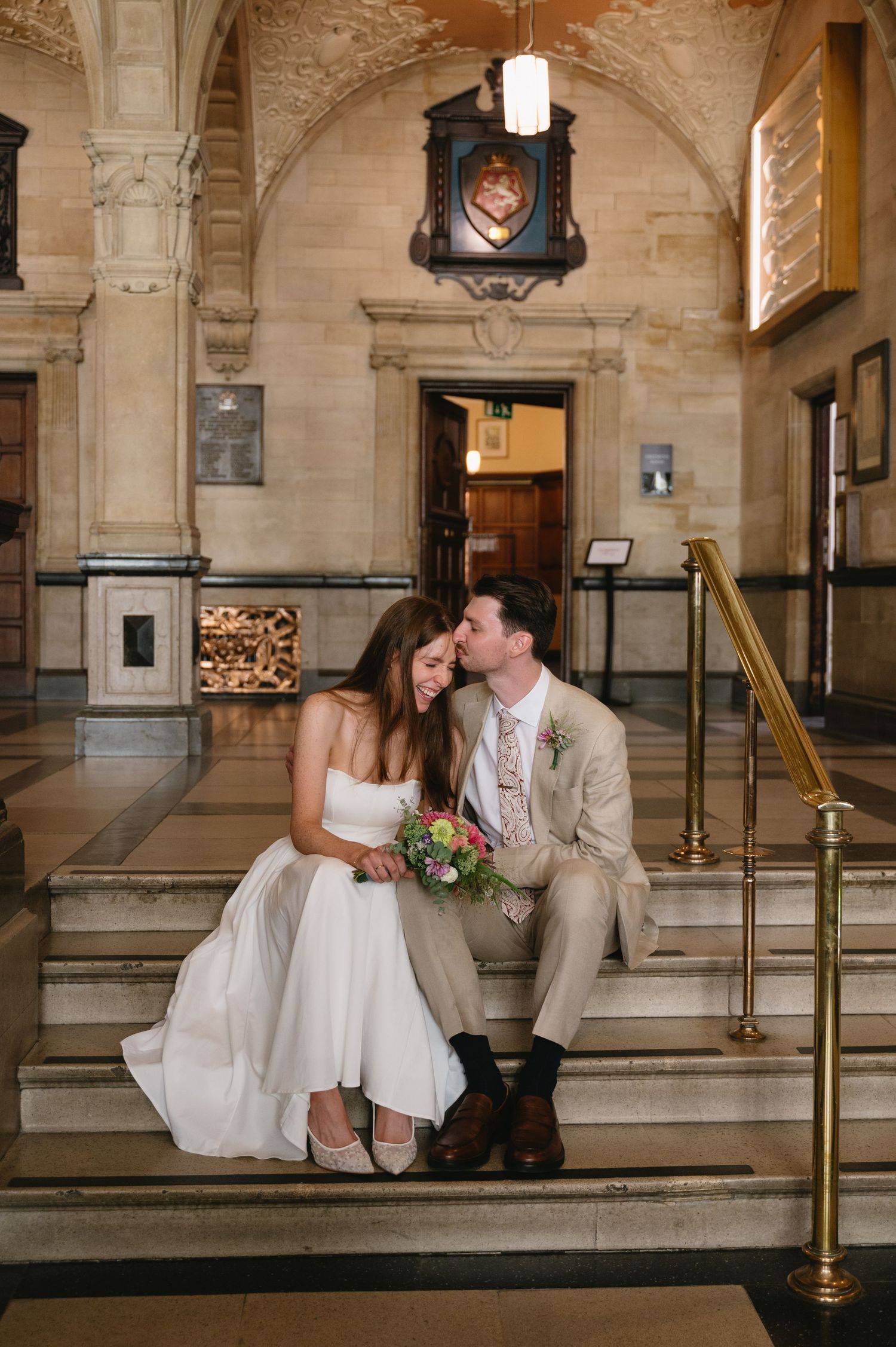 Bride and groom sit intimately on stone steps inside grand historic wedding venue.