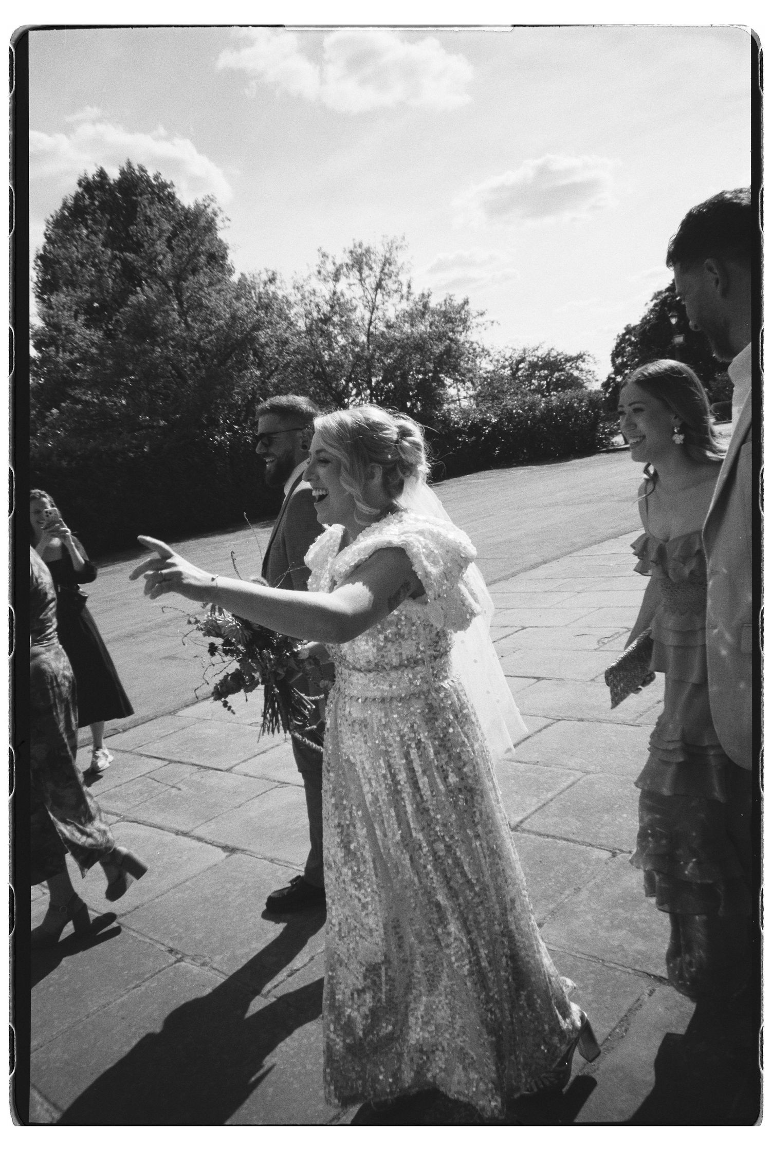 Black and white photo of a bride in lace dress and veil walking outdoors with wedding guests.