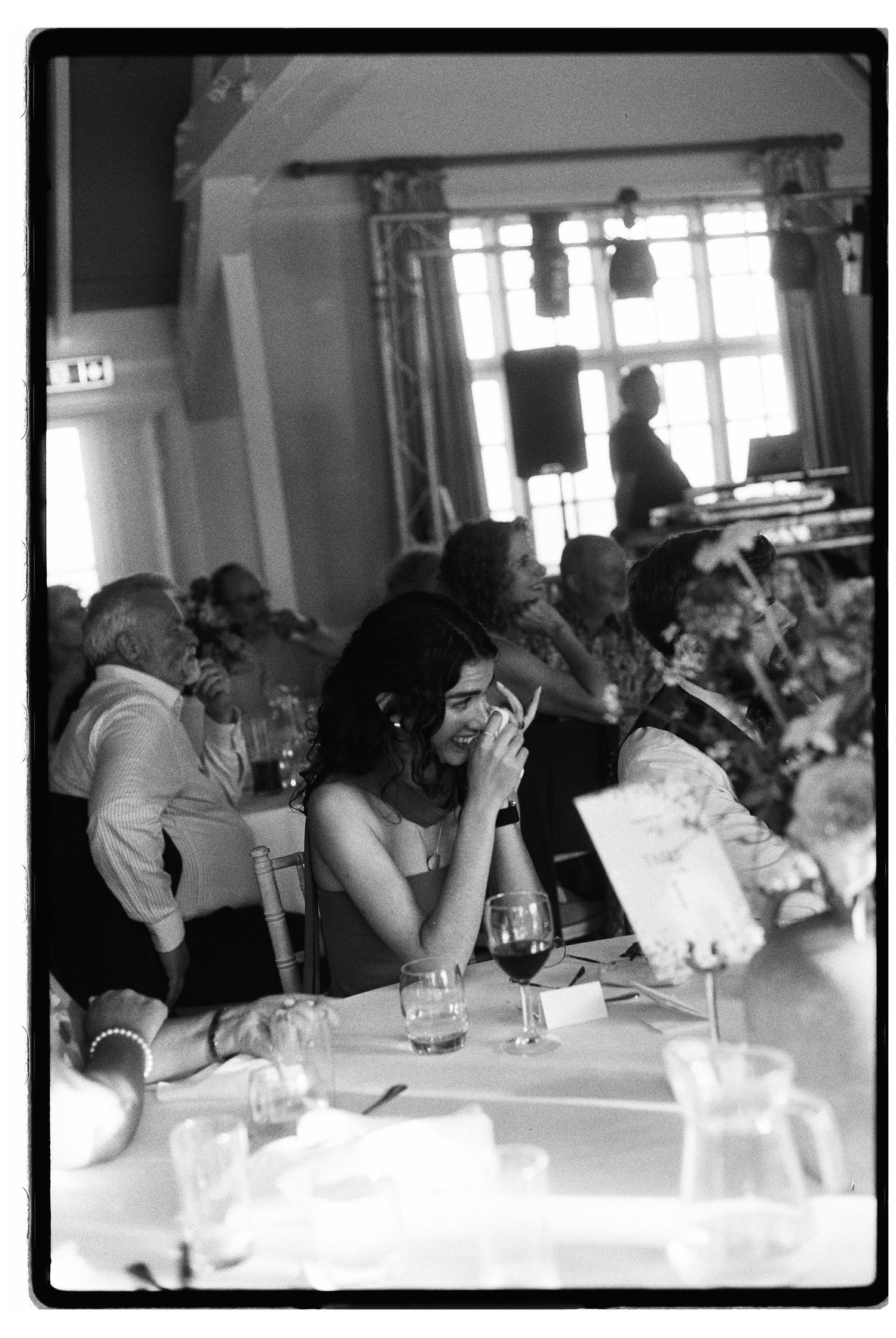 Wedding reception guests seated at tables laughing and socializing in elegant venue, captured in black and white film photography.