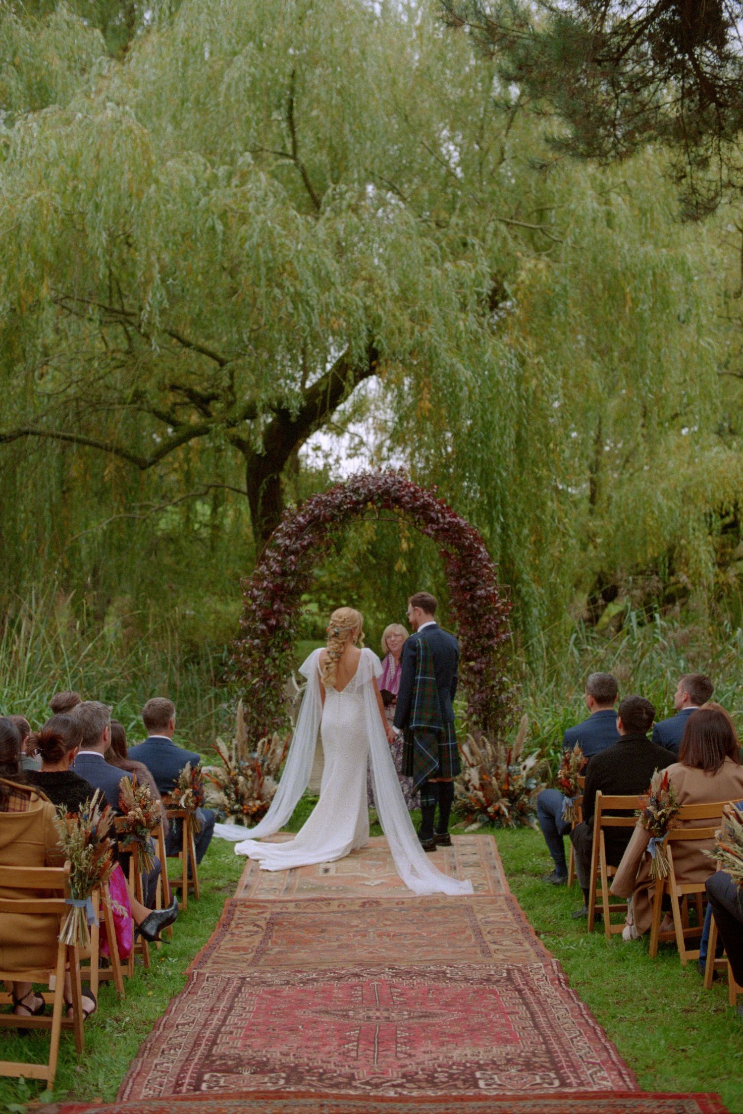Outdoor wedding ceremony under a weeping willow tree with a dark floral circular arch.