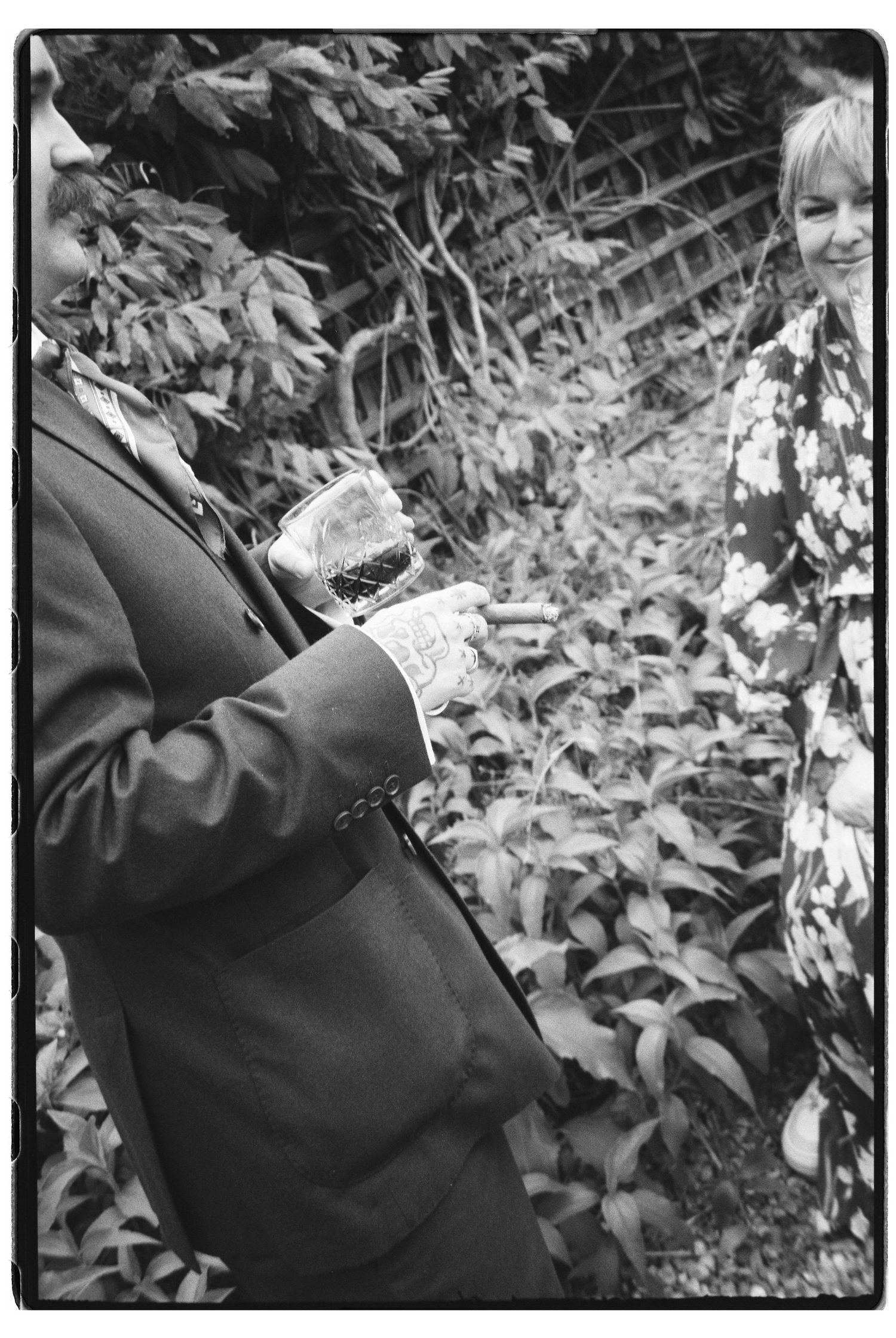 Wedding guest in a suit holds a drink while chatting outdoors among lush green foliage in black and white.