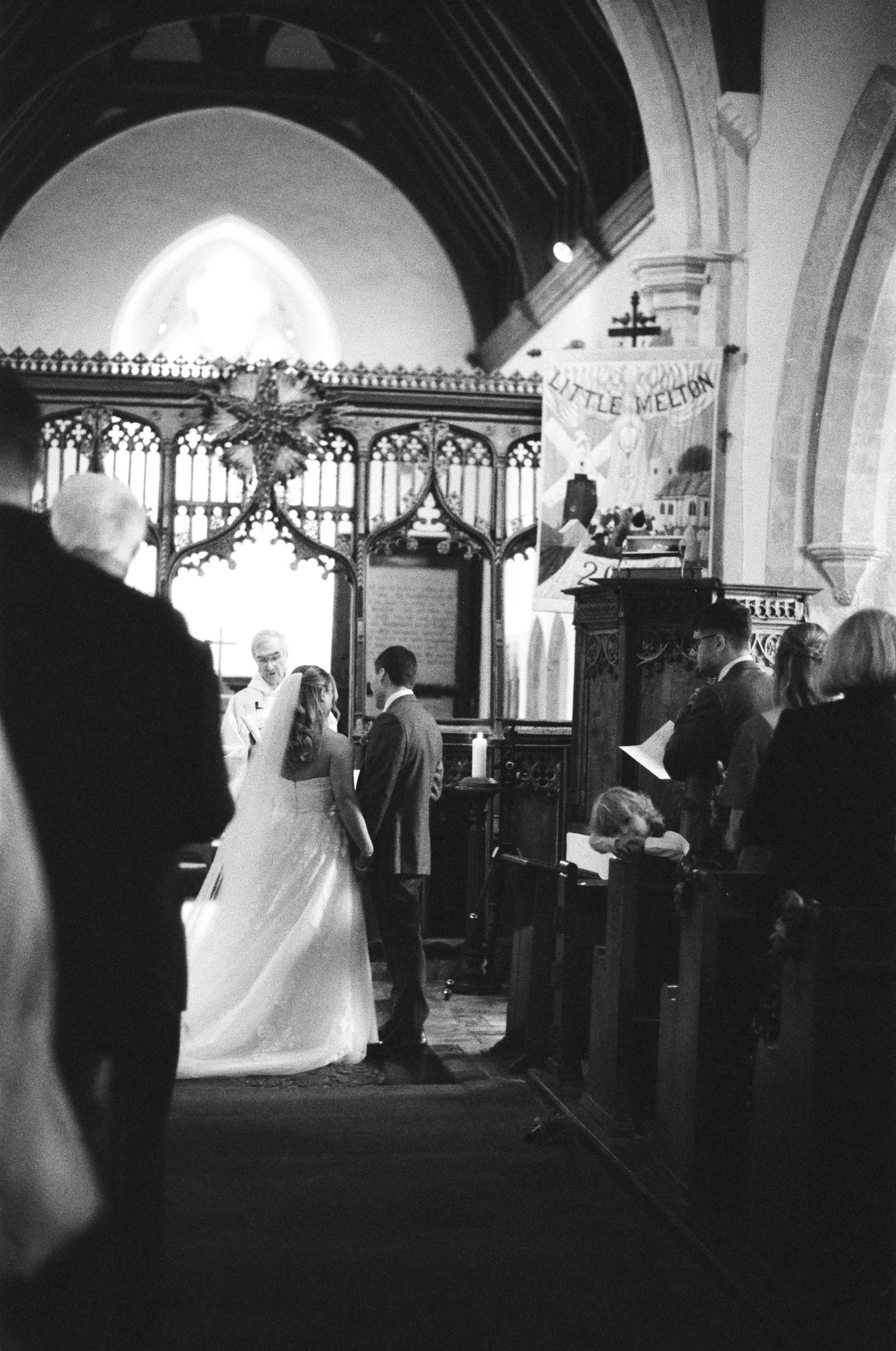 Black and white photo of bride and groom exchanging vows in historic Little Melton church interior.