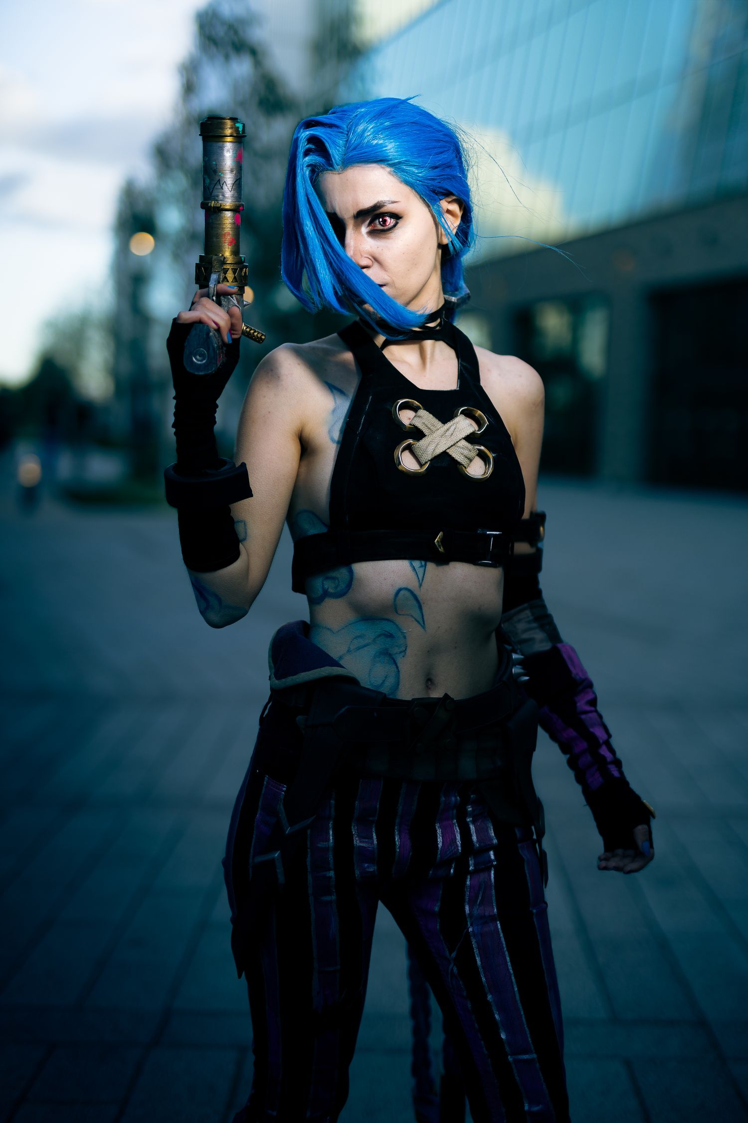Jinx cosplay with bright blue hair poses with a weapon, staring intensely in an urban setting.