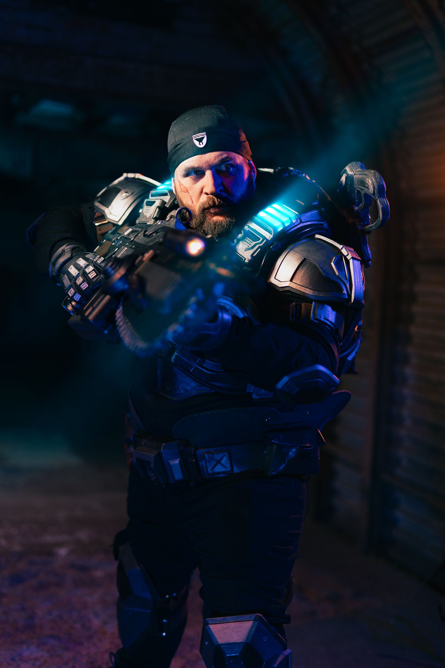Man in blue sci-fi armor cosplay aiming glowing futuristic weapon in dramatic dark setting.