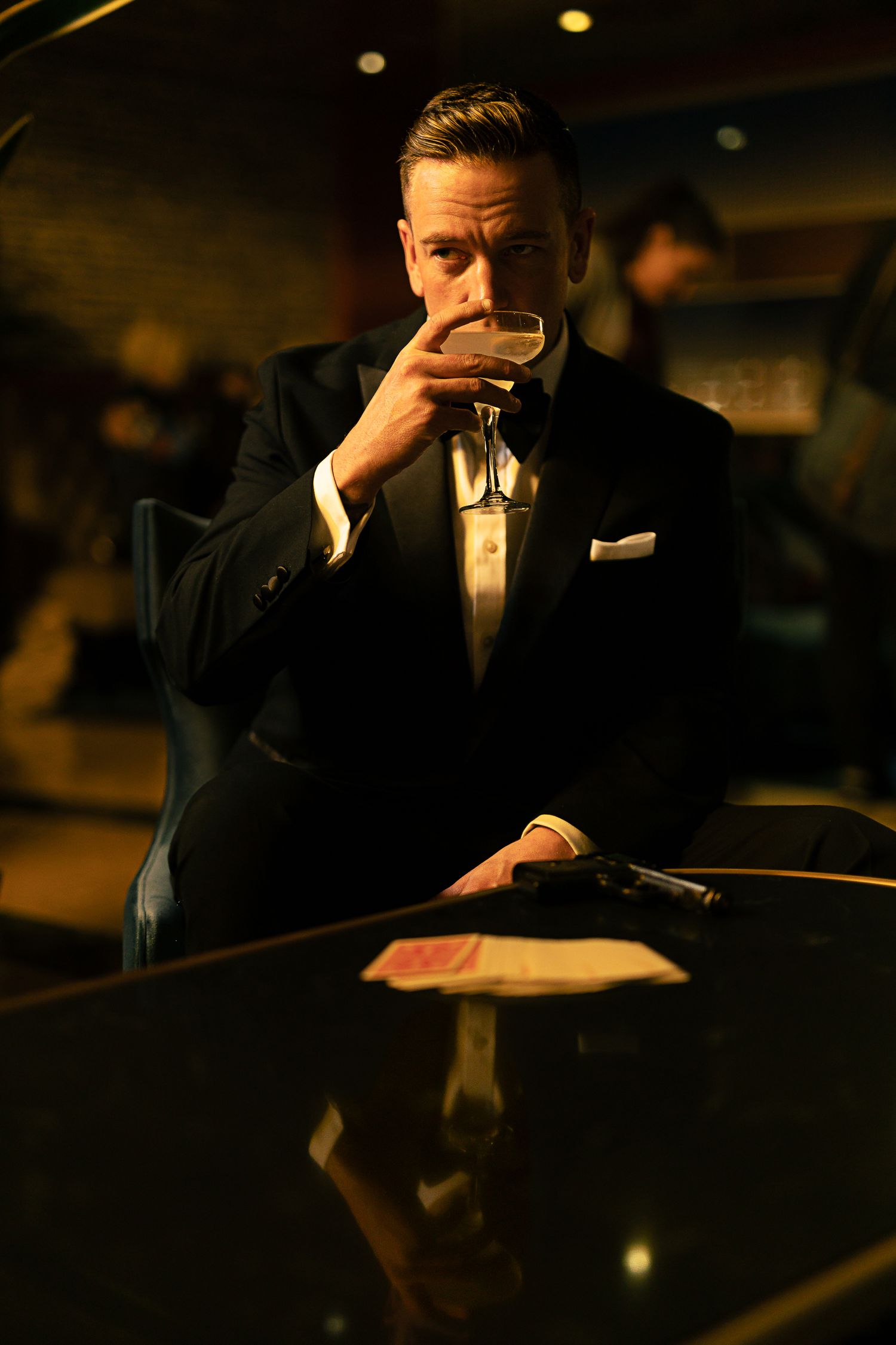 James Bond cosplay in a tuxedo sips a martini at a casino table, cards and a pistol in the foreground.