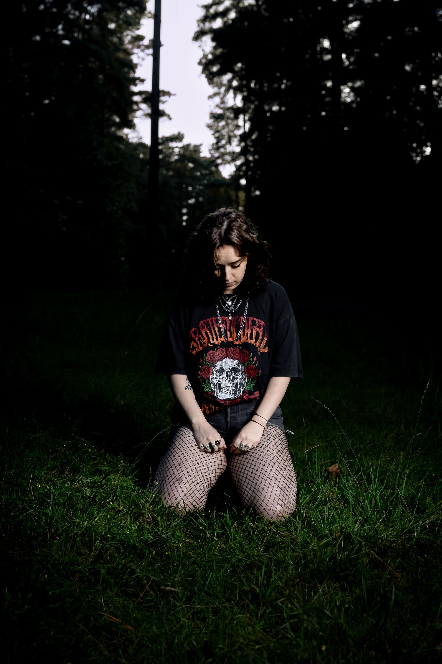 Dark woodland scene with figure seated in grass wearing alternative style clothing.
