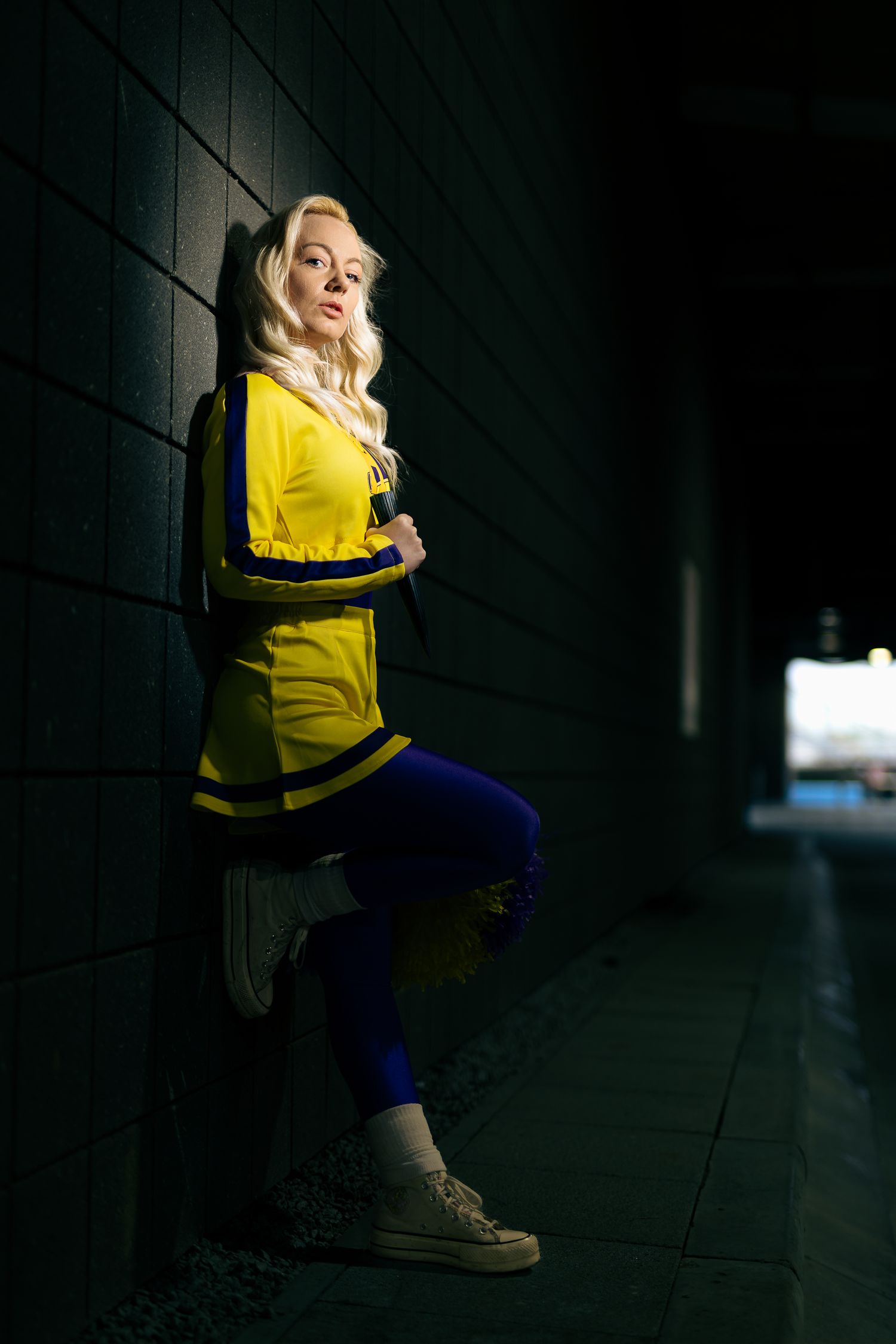 Cosplayer in yellow and blue outfit poses dramatically against dark wall.