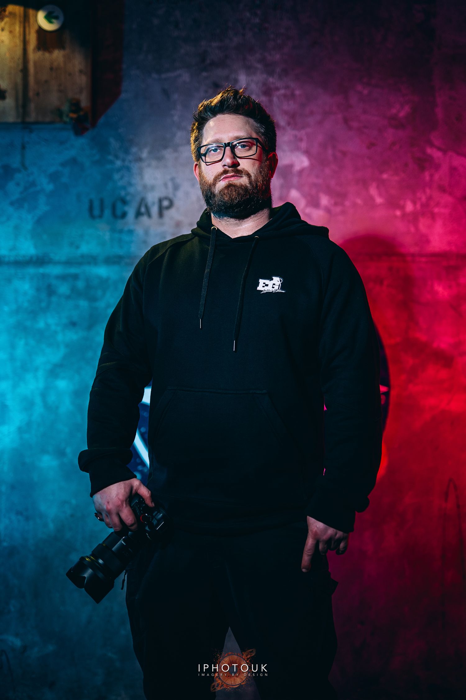 A bearded man in a black hoodie holds a camera, posed against a dramatic blue and red lit grunge wall backdrop.