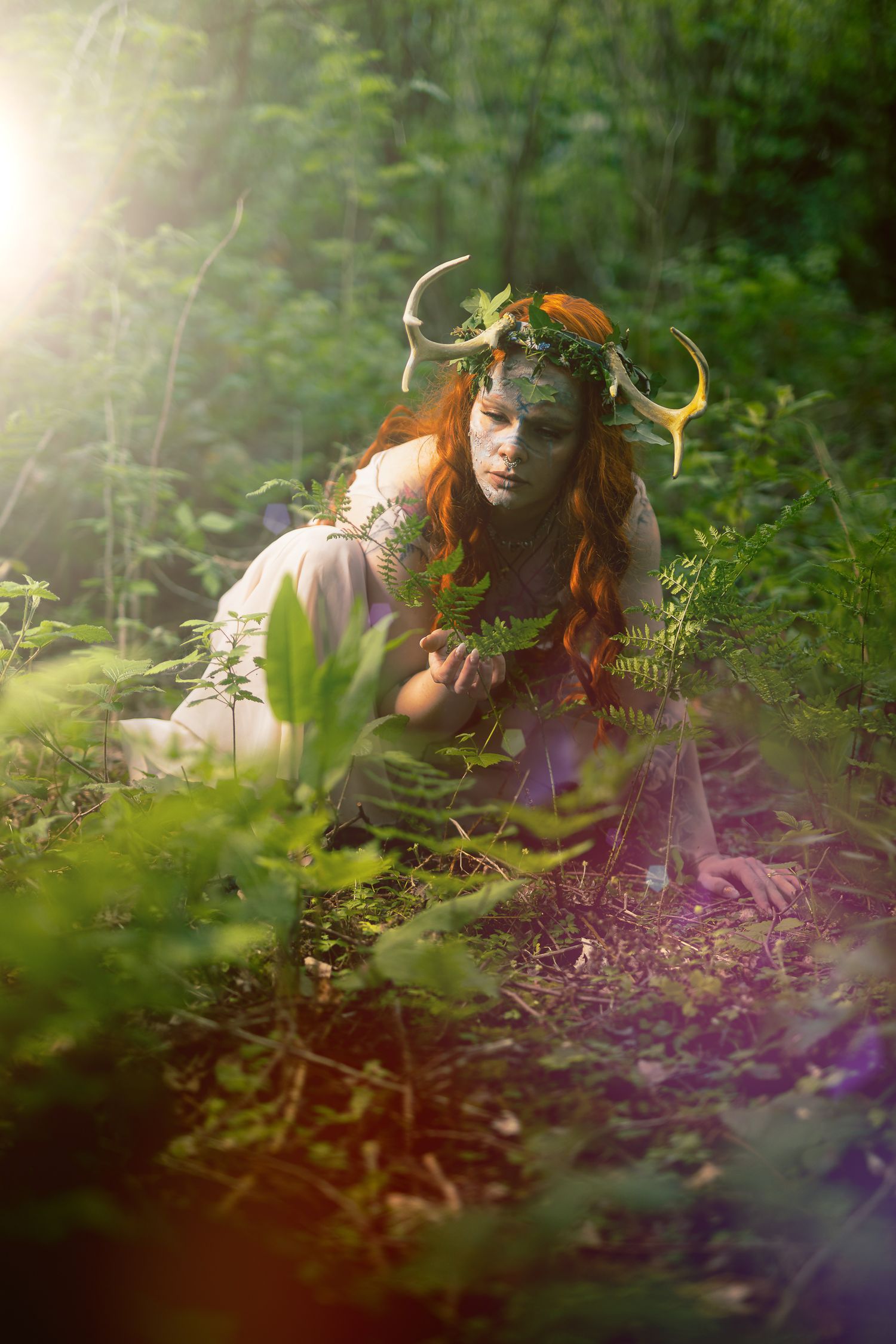 Fantasy forest creature with antlers, floral crown, and body paint crouching among lush green plants in sunlit woods.