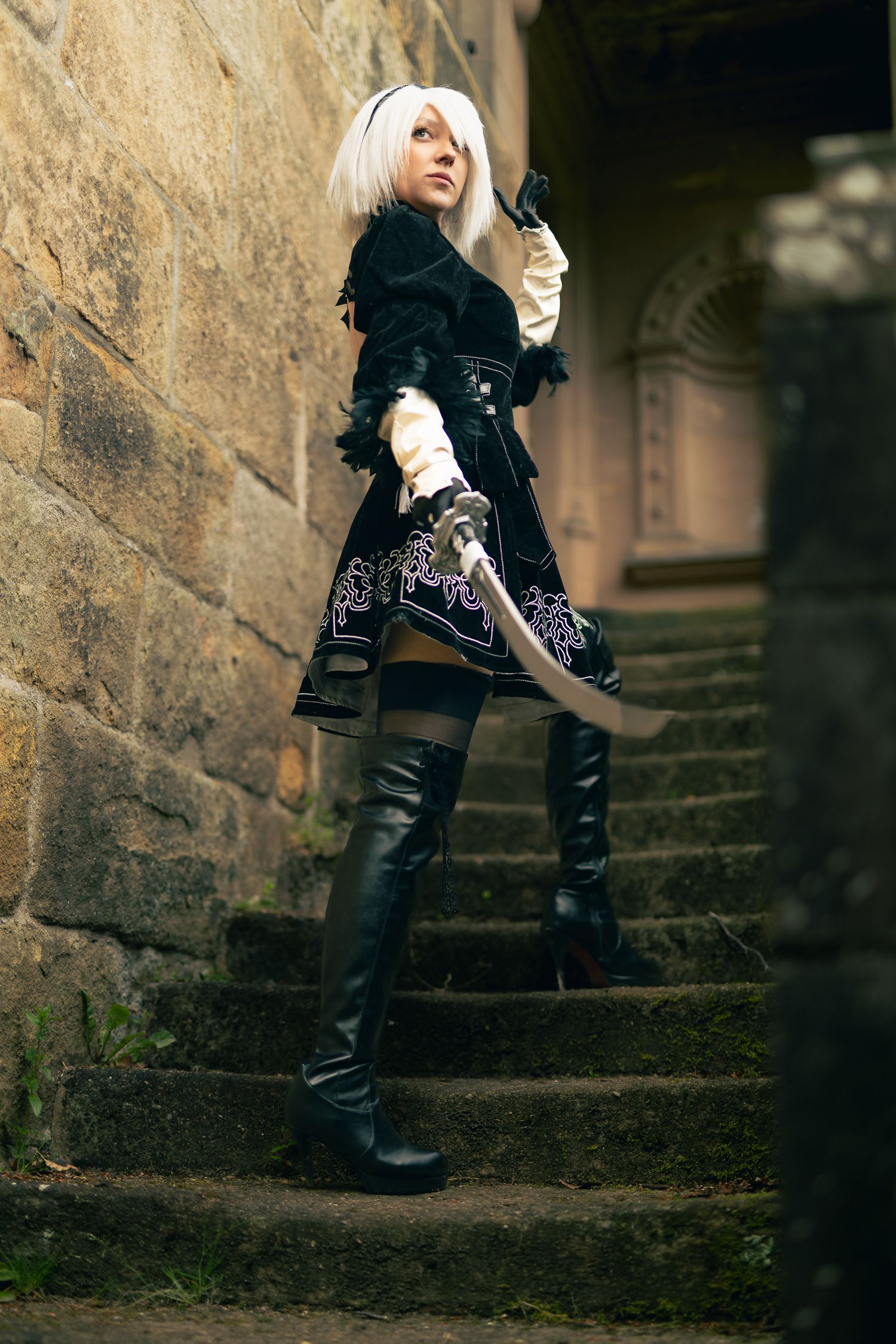 Person cosplaying as 2B from NieR: Automata, wearing black dress and boots, holding a sword on stone steps.
