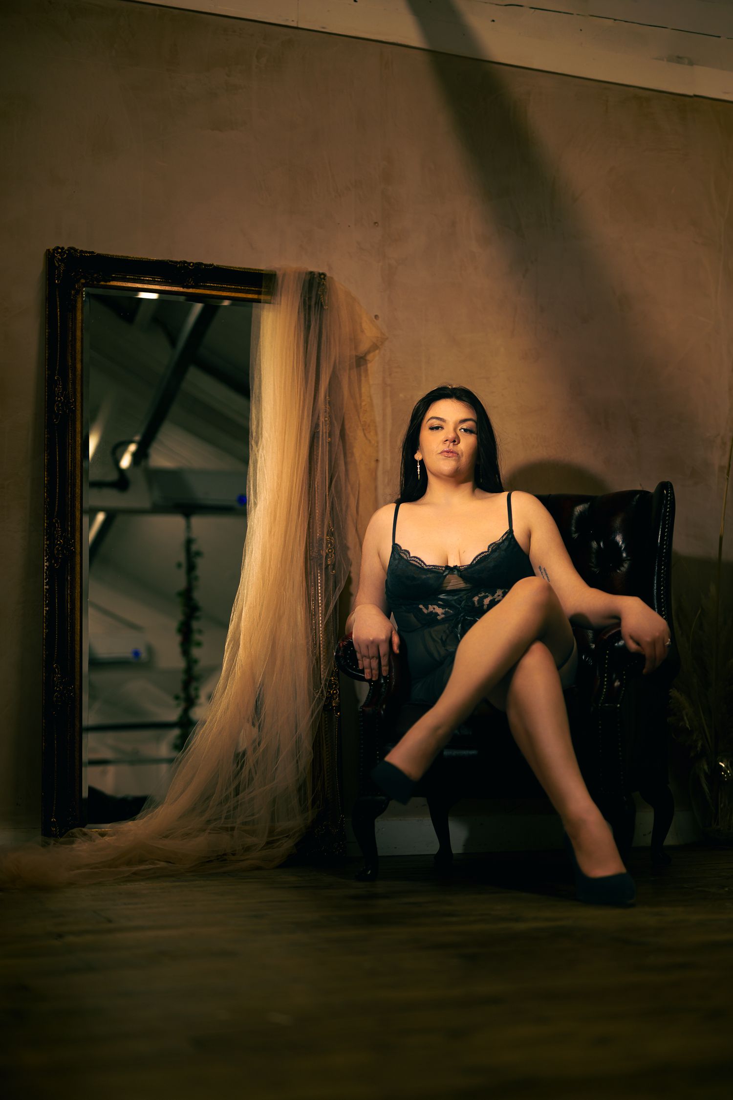 Boudoir portrait of a model seated in a leather armchair beside an ornate mirror draped with sheer fabric