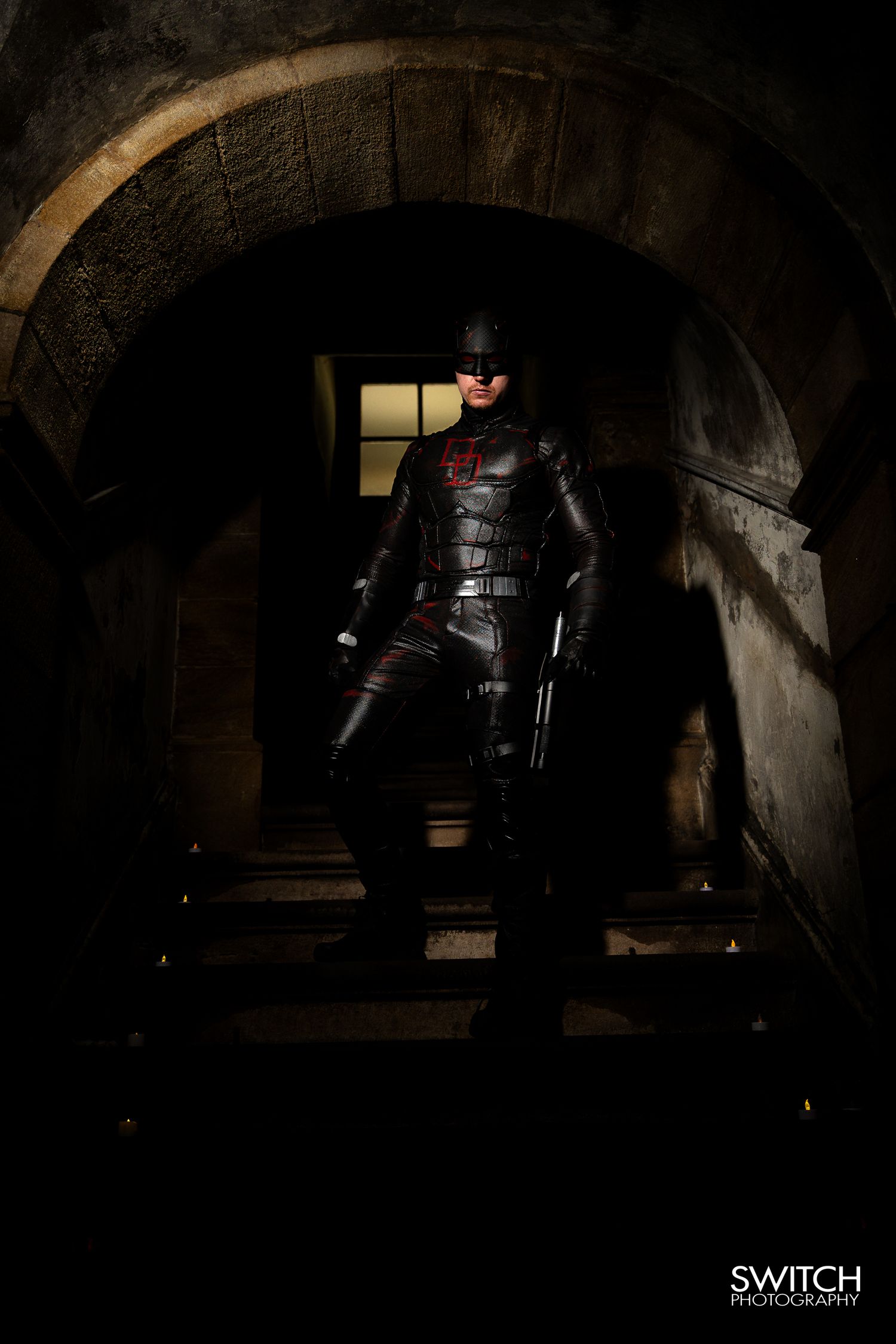 Daredevil cosplay under a stone archway, dramatic low-key lighting with deep shadows and a moody backdrop.