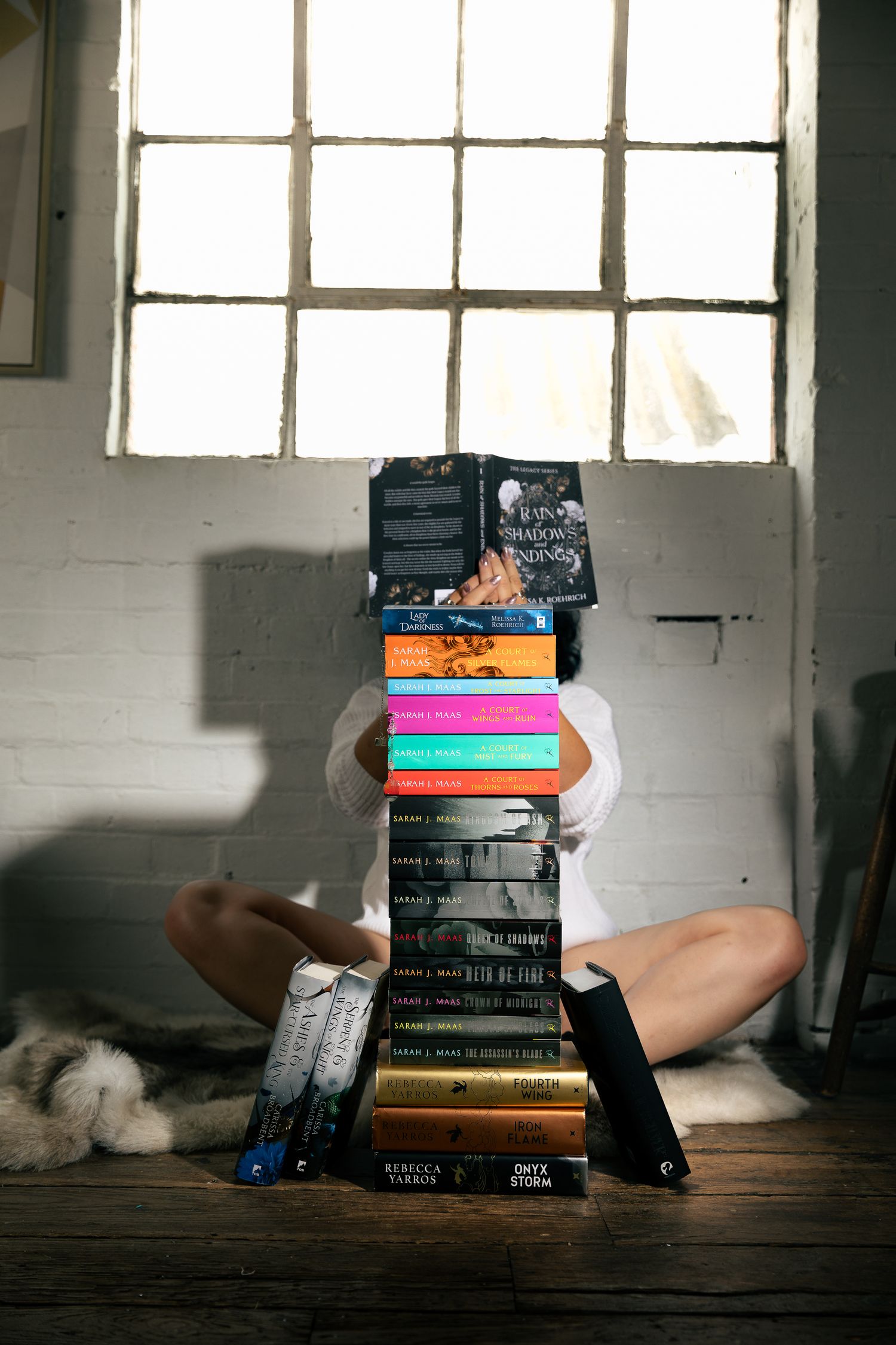 Stack of books arranged on a wooden floor near a bright window in an industrial space.