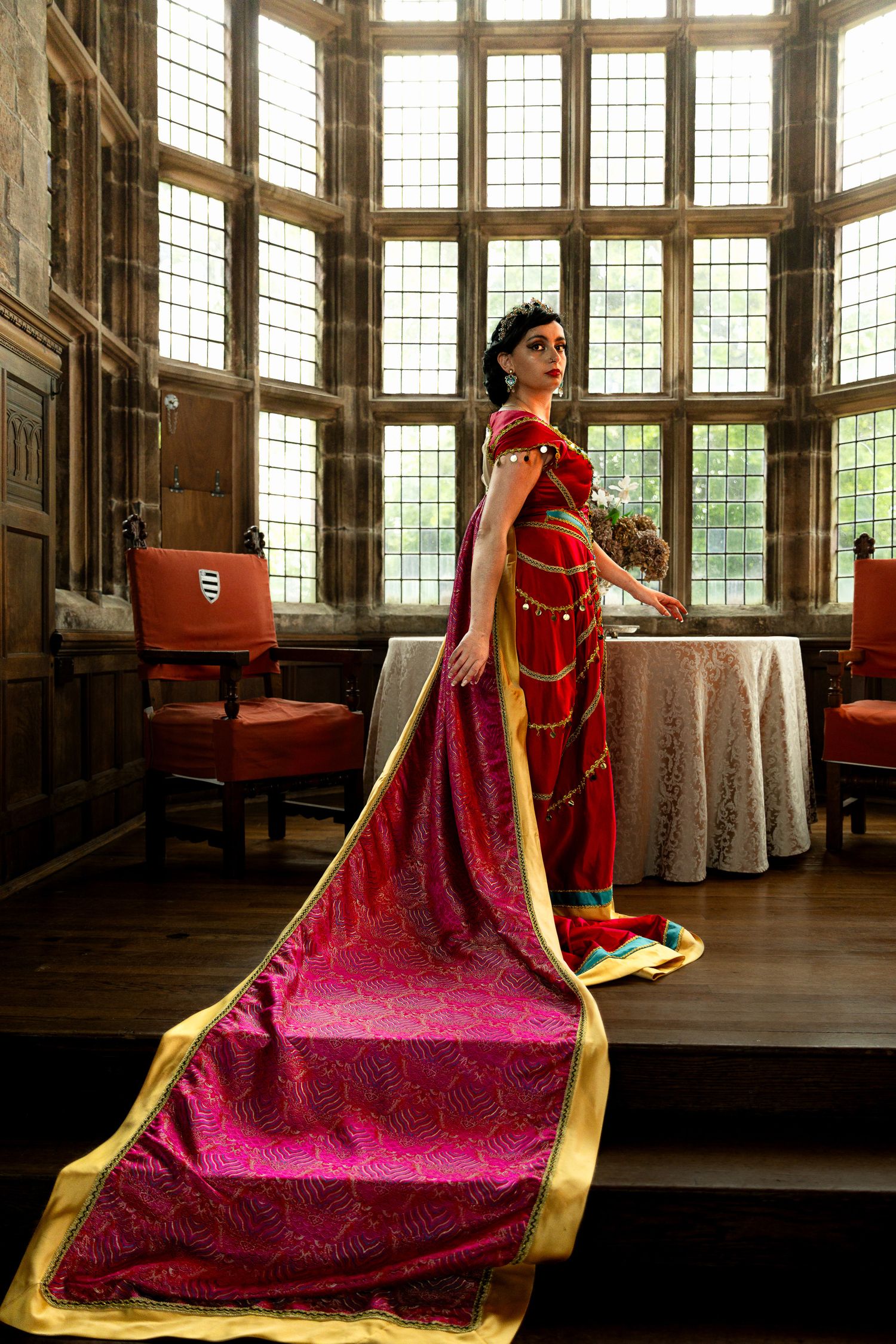 Elegant cosplay in ornate room featuring flowing red and gold royal gown.