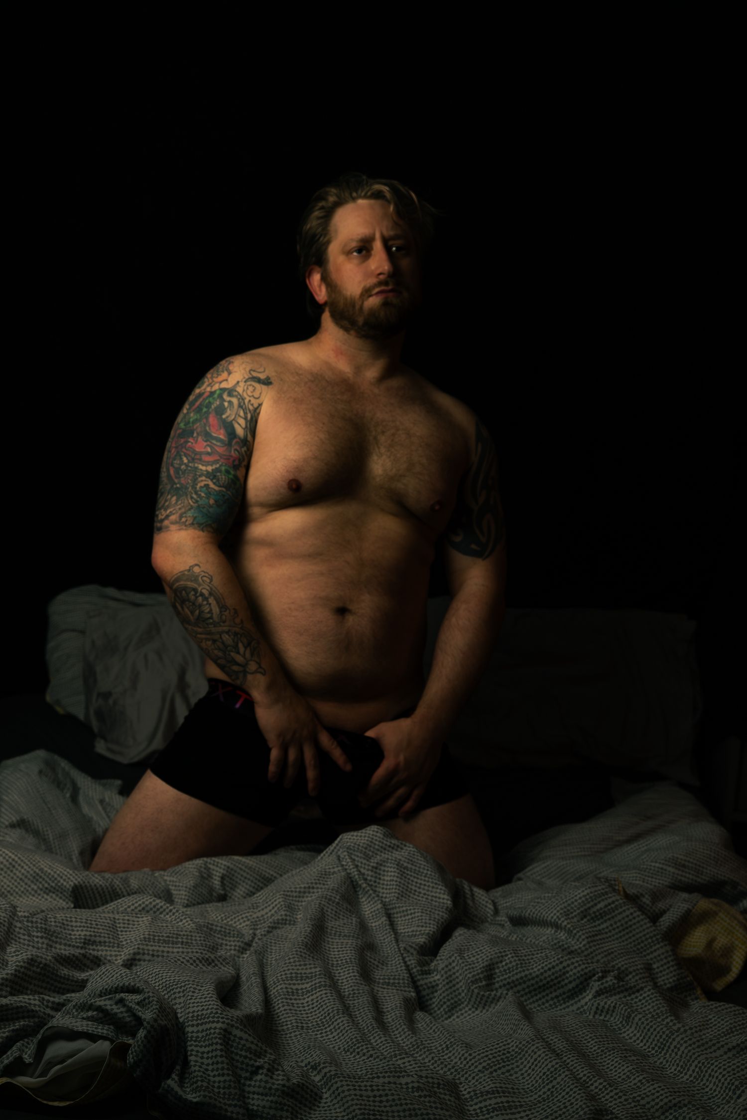Moody bedroom photoshoot with dramatic lighting highlighting tattoo art.