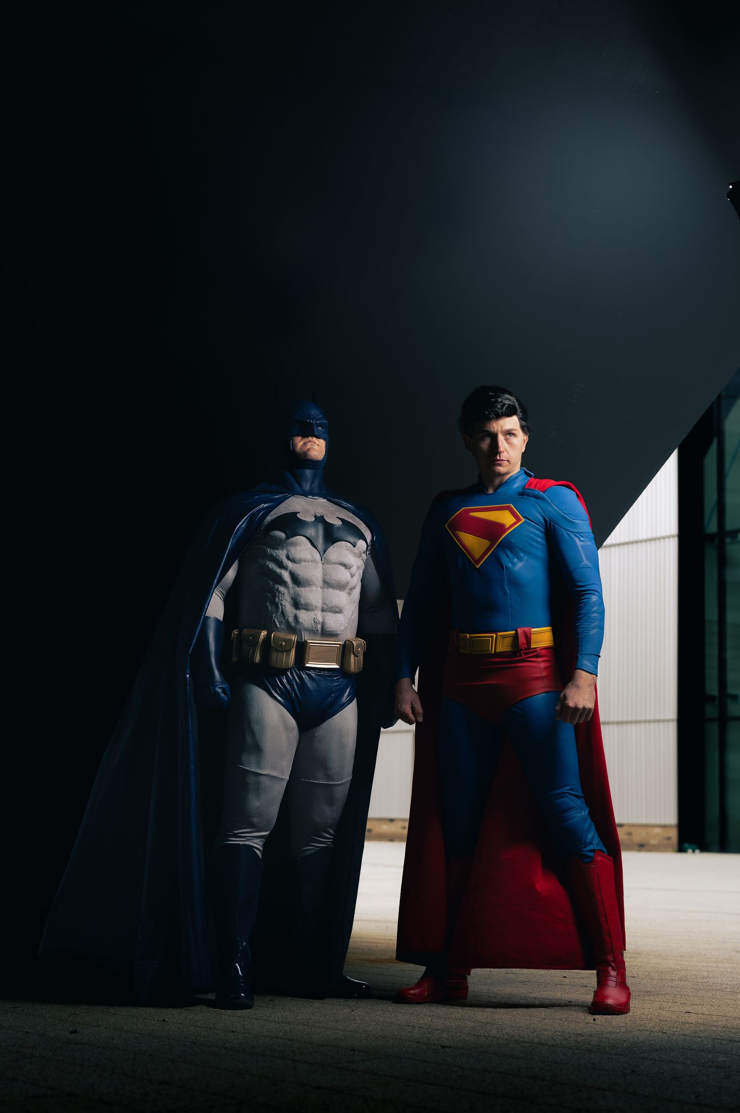 Batman and Superman cosplayers stand side by side beneath a dark structure, framed by dramatic negative space.