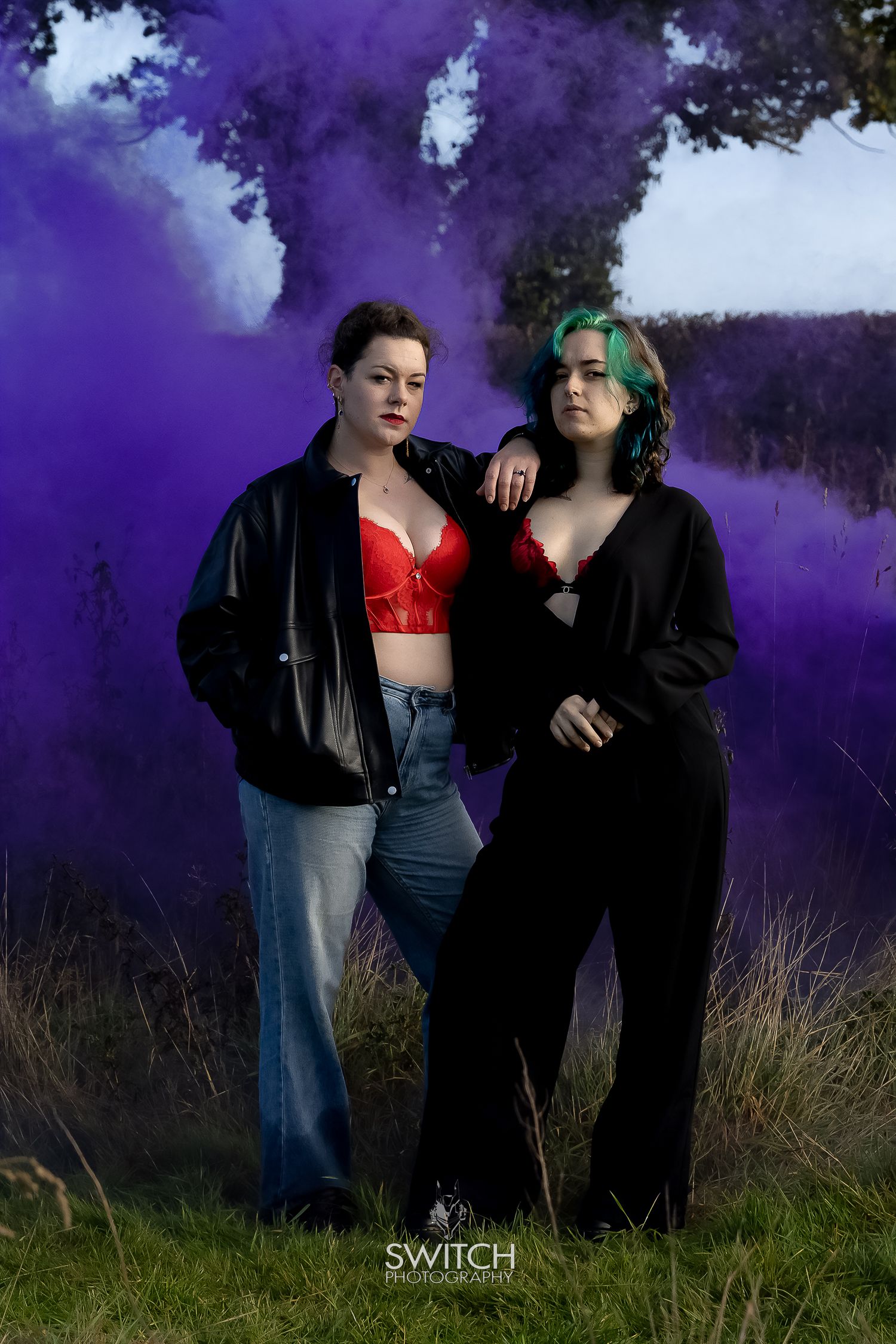 Two women posing in a field with purple smoke behind them, wearing dark outfits with red accents and strong, confident expressions.