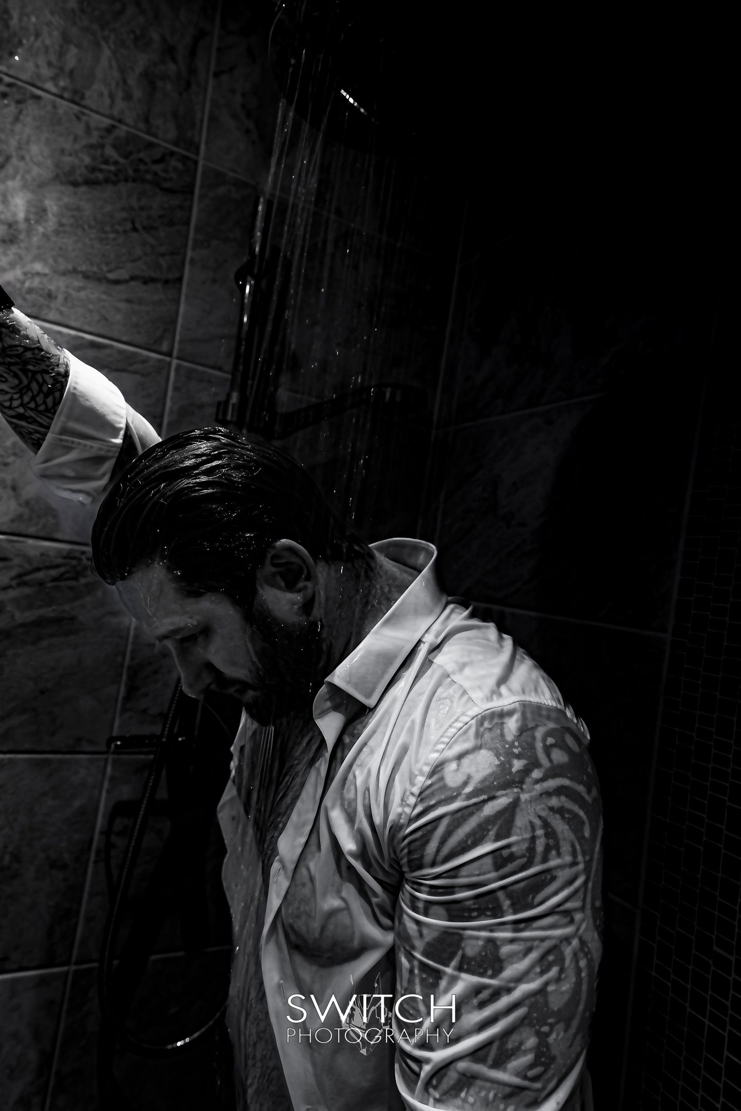 Black-and-white portrait of a tattooed model under a shower, wet shirt, head lowered as water falls.