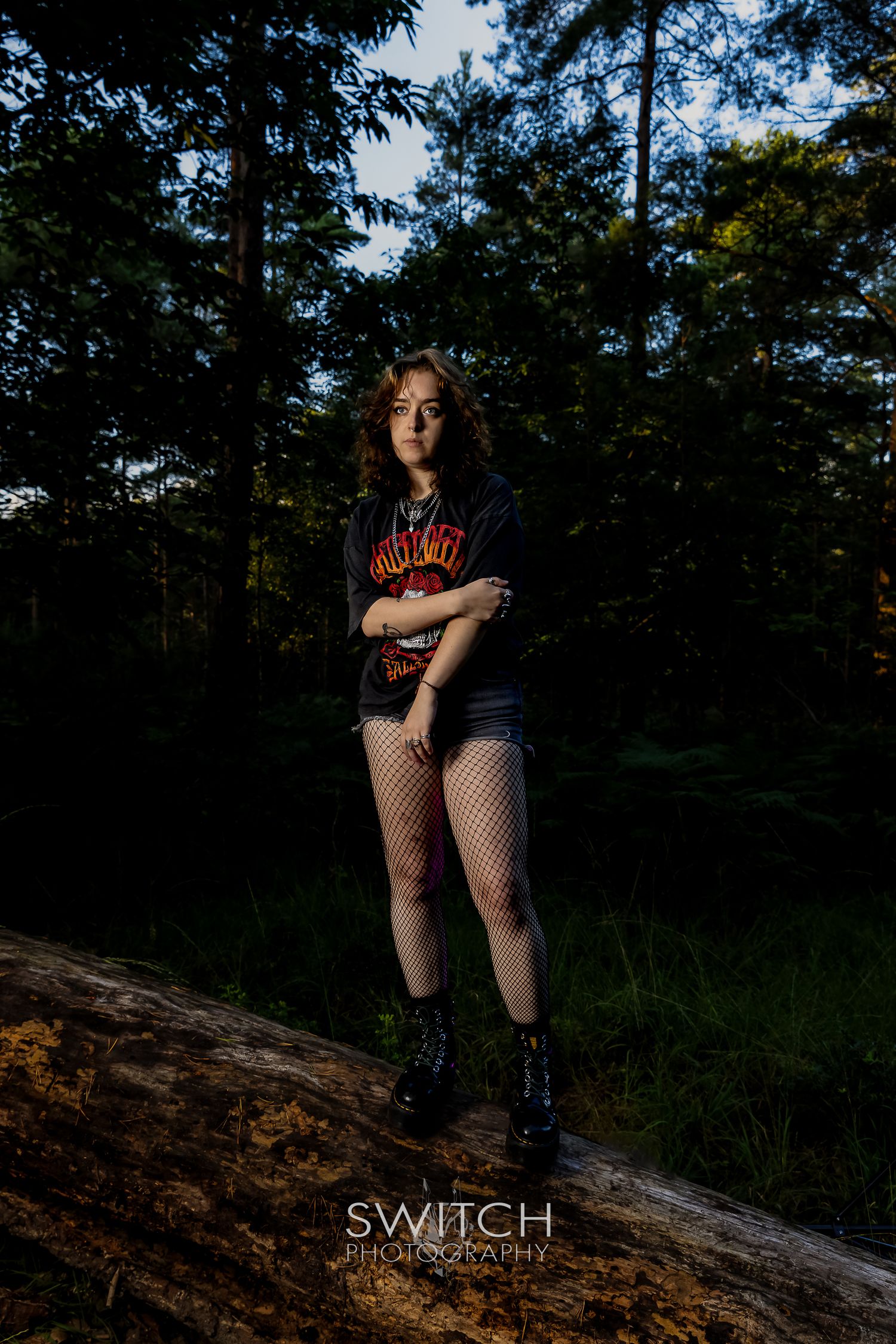 Outdoor portrait of a woman standing on a fallen log in a forest, wearing a dark graphic tee and fishnet tights in dramatic light.