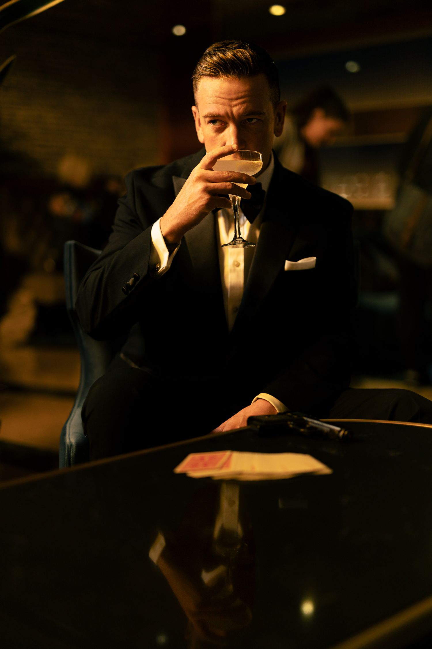 A suave man in a black tuxedo sips a drink at a bar with a gun on the table in a dimly lit setting.