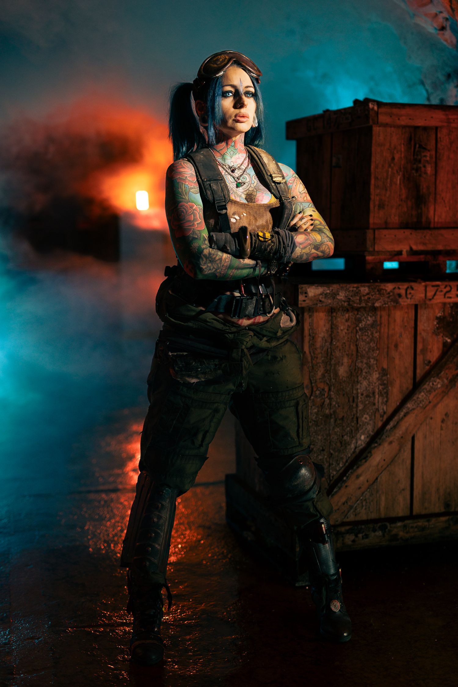 Tattooed woman in tactical gear poses arms crossed in a smoky, dramatic setting with orange and teal lighting.