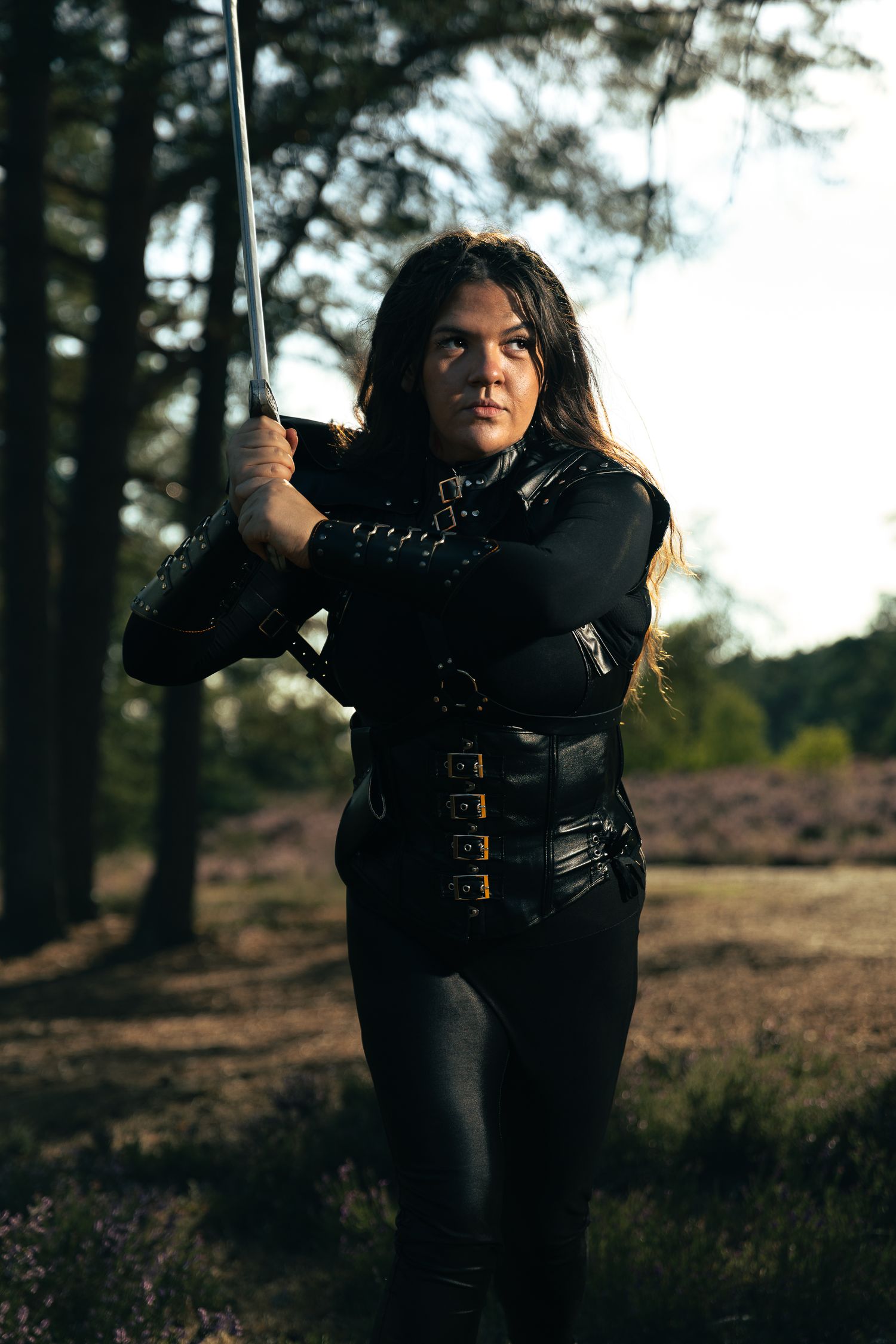Nesta Archeron cosplay in black leather armour, grips a raised sword in sunlit woodland.