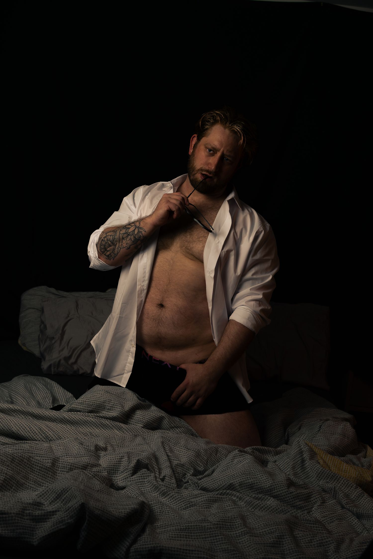 Intimate portrait series in white shirt on dark bedding with dramatic shadows.